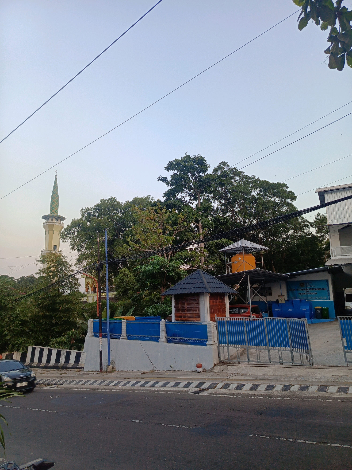 The image shows a street scene in Magetan, Indonesia, with a mosque minaret visible in the background. The foreground features a road with a black car driving by. To the right of the road is a sidewalk and a low wall topped with a blue fence. Behind the fence is a building with a blue sign that appears to be a government institution, possibly related to health or nutrition, with text in Indonesian. There are also several large blue water tanks and a small, dark-roofed structure. Dense green trees fill the middle ground, obscuring much of the view behind the fence. Power lines are visible crisscrossing the sky. The overall lighting suggests it is either early morning or late afternoon.