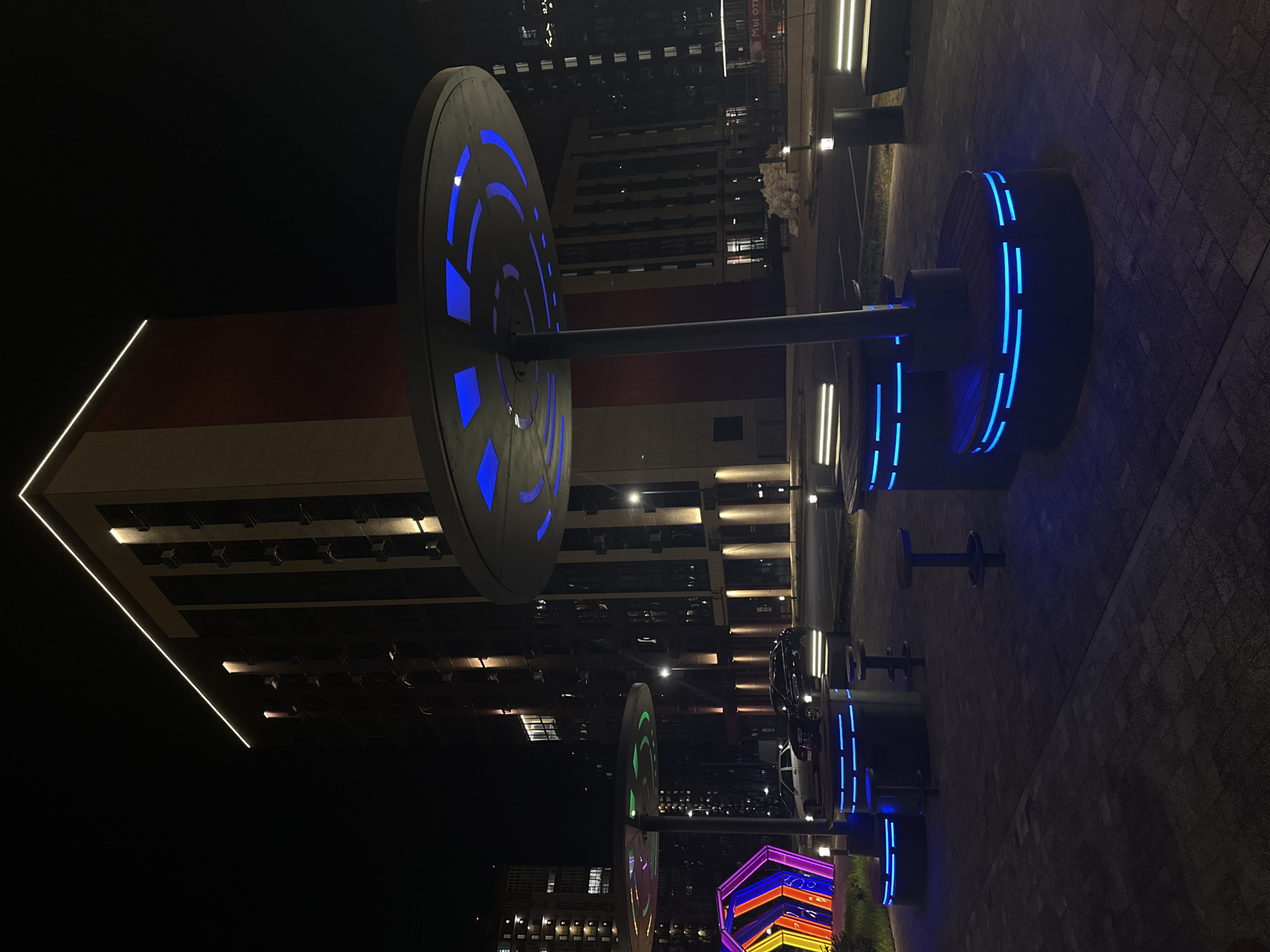 At night, the city of Rostov-on-Don, Russia, shows a modern outdoor social area. The ground is paved with gray bricks. This space features several circular, illuminated benches with individual stool seating. The benches have horizontal blue neon bands around their circumference. The stools are a plain dark color. Rising above the benches are decorative metal discs on poles, which also have integrated blue lighting. A tall, light-colored building with a red section, and a bright white trim at the top, is visible in the background. There are other modern buildings in the area and some of the windows have lights on. A colorful, geometrical light installation can be seen toward the bottom of the shot. There are no people visible. The overall impression is of a contemporary, illuminated urban space.