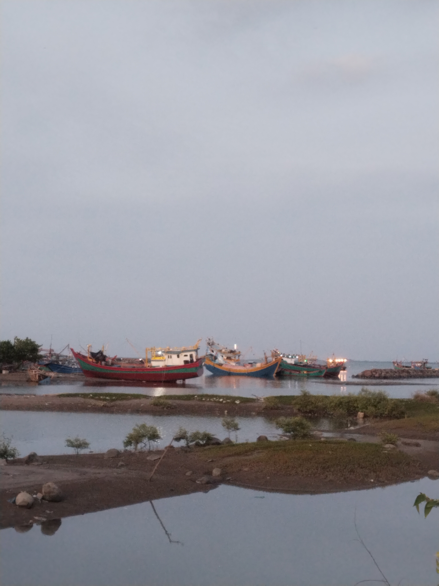 The image captures a waterfront scene in Lhokseumawe, Indonesia. Several fishing boats of varying colors and sizes are docked or grounded in the shallow water near the shore. The foreground shows a muddy bank with sparse vegetation. The sky is overcast, contributing to a subdued, grayish light over the entire scene. The water appears calm, reflecting the sky and the boats. No people are visible in the frame. The overall setting suggests a fishing community or a docking area for fishing vessels.