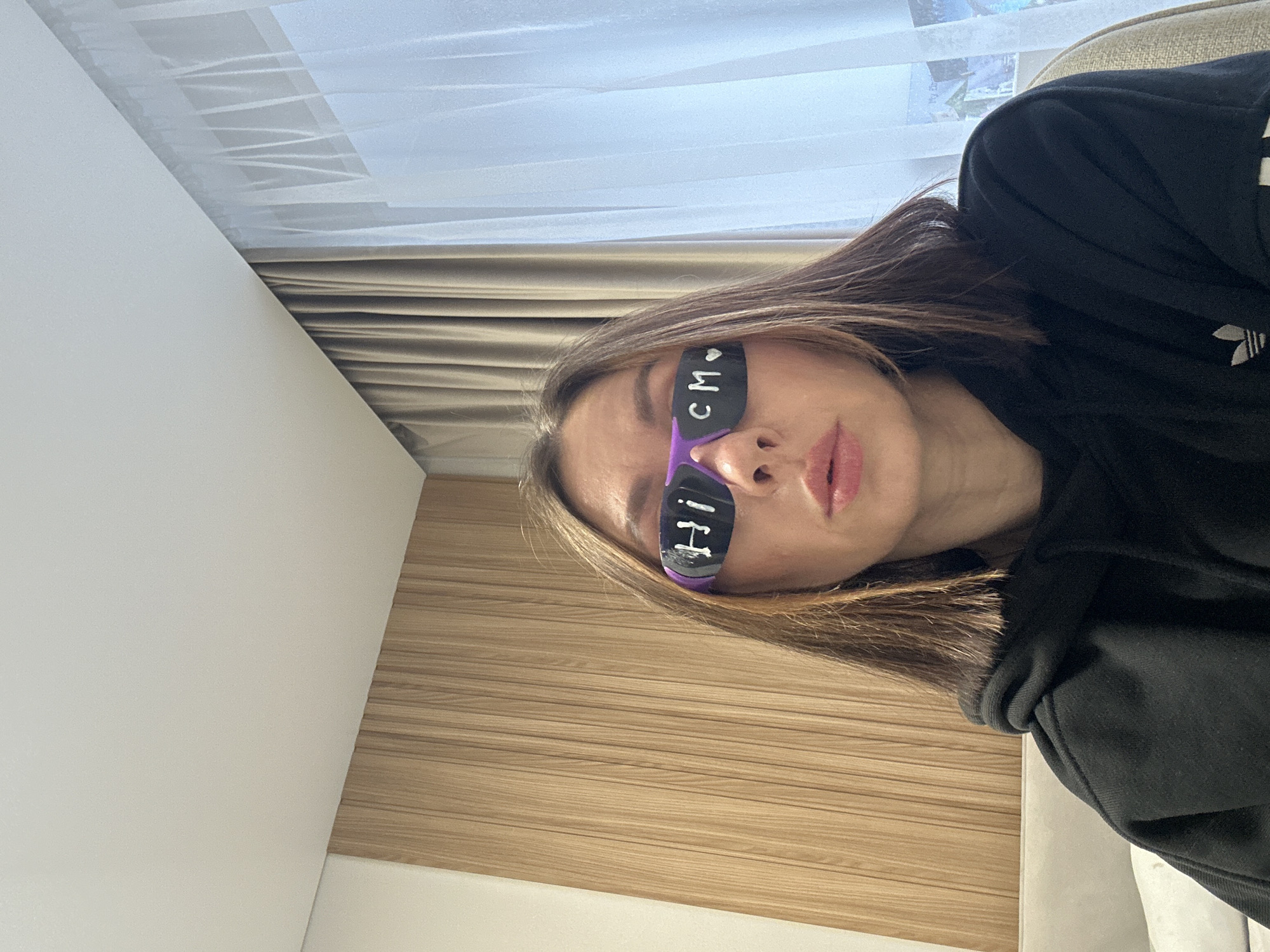 A woman wearing purple-framed sunglasses with the words "HI" and "CM" written in white marker on each lens is posing for a selfie. She is wearing a black Adidas hoodie. The background consists of a white wall to the left, light wood paneling on the bottom right, and a window covered with a white sheer curtain and a beige thicker curtain. The location is indoors but there is no information provided to establish the city of Votkinsk, Russia.