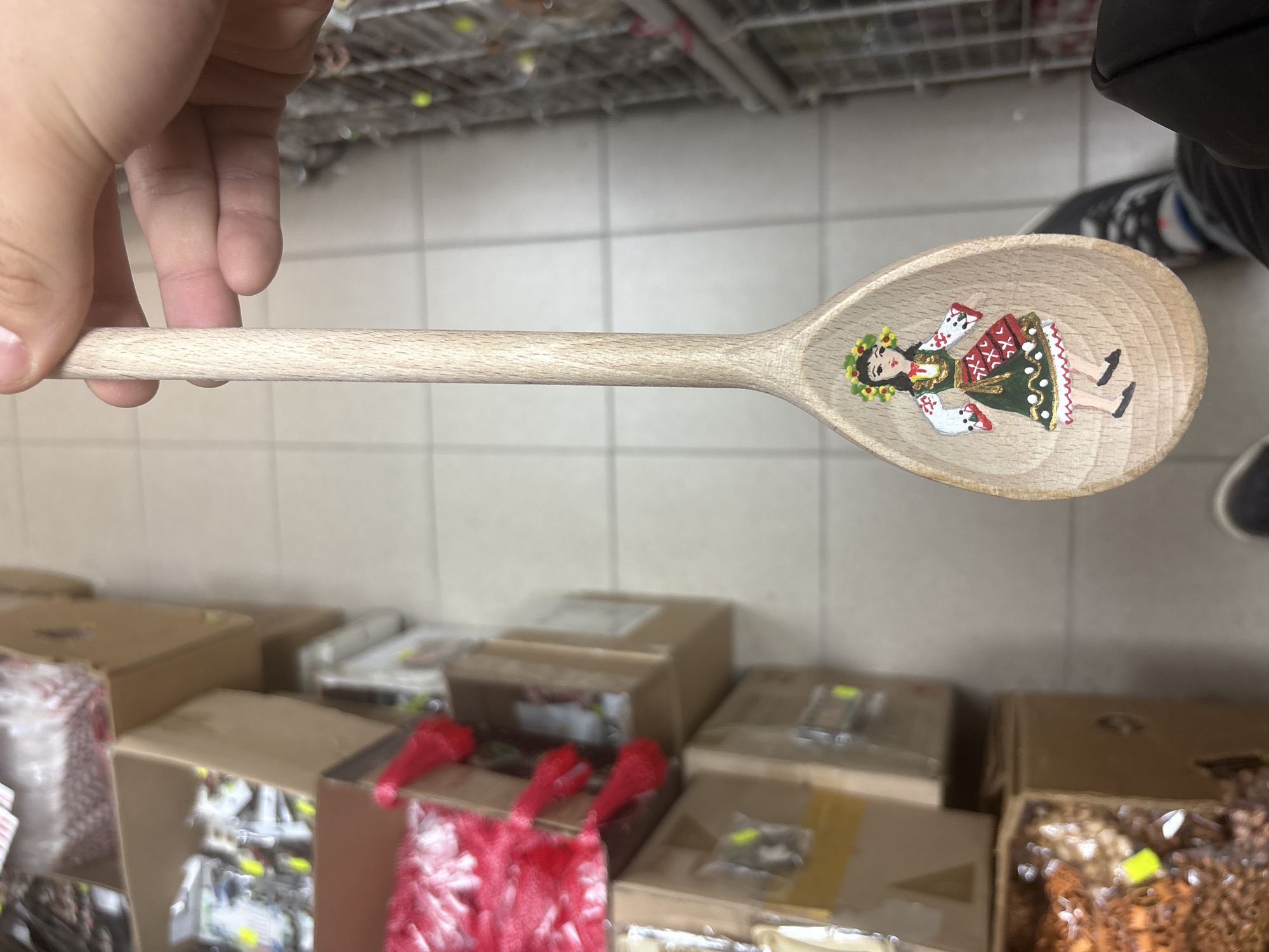 A person is holding a wooden spoon in their left hand. Inside the spoon is a small painted figure of a person wearing traditional Bulgarian clothing. The background shows shelves and boxes filled with merchandise, suggesting a retail setting. Based on the painted figure of a person wearing traditional Bulgarian clothing on the wooden spoon, the location is likely Sofia, Bulgaria.