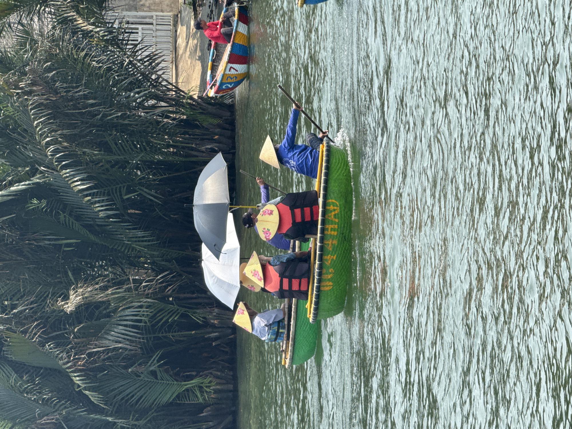 This vibrant daytime photograph depicts a group of people enjoying a boat ride on a calm river, framed by lush tropical vegetation.
The primary focus is a long, green boat, likely a traditional Vietnamese basket boat (thúng chai), carrying at least three passengers and one paddler. The paddler, dressed in a blue shirt and a conical hat (nón lá), actively propels the boat using a long pole. The passengers are also wearing conical hats and orange life vests, and two open white umbrellas provide shade from the sun, suggesting a leisure or tourist activity.
In the background, another colorful boat with the numbers "3" and "2" painted on its side is visible closer to the river bank, with one person in a red shirt. The river water has gentle ripples, reflecting the bright daylight. The banks are densely covered with dark green palm trees and fronds, indicating a tropical or subtropical environment. A building with white railings is partially visible behind the foliage on the bank.
The bright daylight and use of umbrellas confirm it is a sunny day. The conical hats and boat styles strongly point to a Vietnamese setting. Yellow text, "ĐƯỜNG HẠ", is visible on the side of the green boat.
