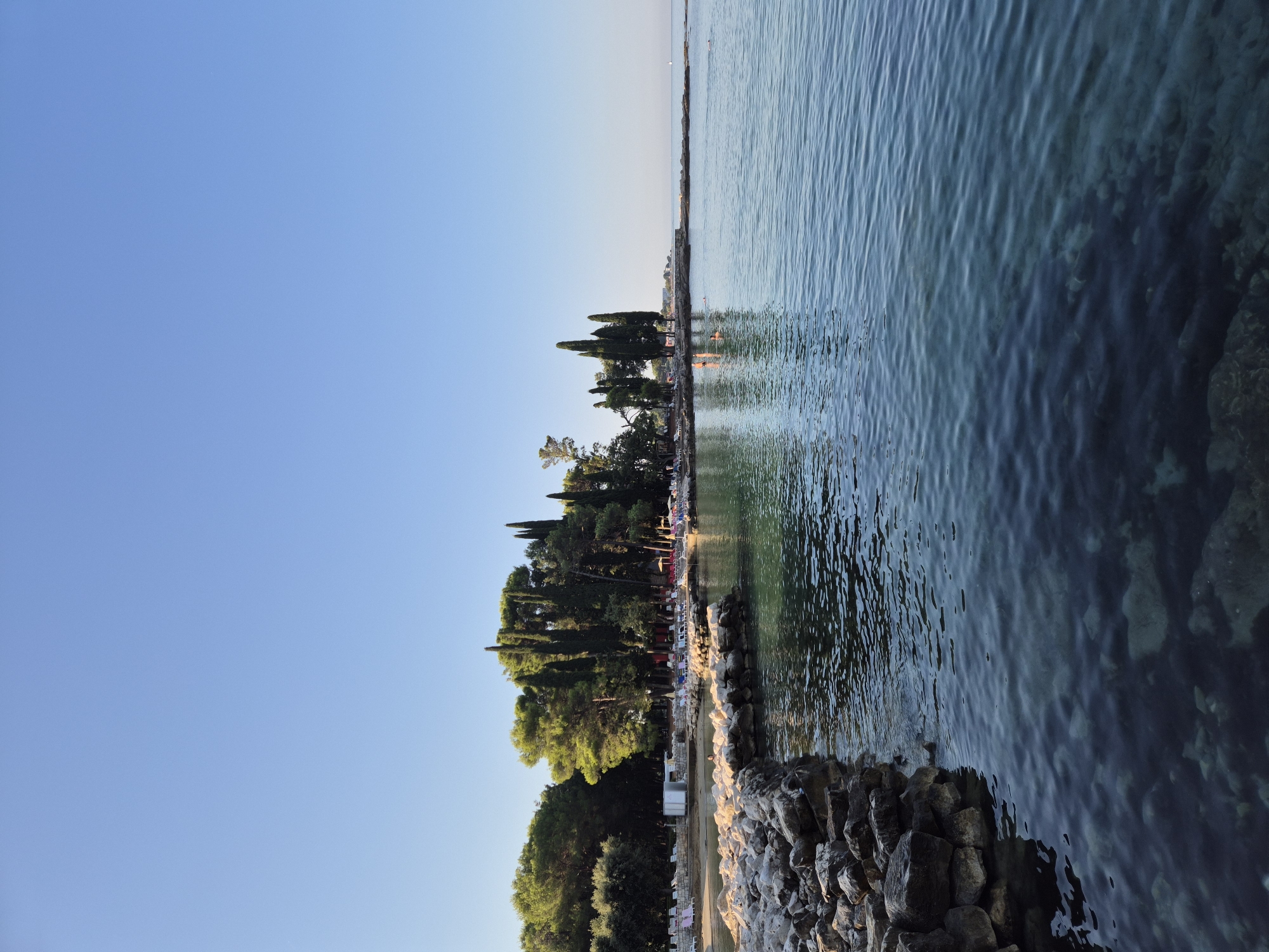 The image depicts a serene coastal scene in Monterol, Croatia. The foreground is dominated by a rocky breakwater extending into the clear, turquoise water of the Adriatic Sea. The water is calm with gentle ripples reflecting the clear blue sky. Submerged rocks are visible beneath the surface, indicating shallow depths.
Beyond the water, a well-maintained beach area is visible, lined with lounge chairs and umbrellas. A cluster of tall, slender cypress trees and other lush green foliage forms a backdrop to the beach, creating a natural barrier. Several people are present in the water, some swimming or wading, while others are visible on the beach. The sun is shining brightly, casting a warm glow over the scene, suggesting it is either early morning or late afternoon. The horizon is clear, with no visible boats or ships, except for a small white sail in the far distance. The overall impression is one of a tranquil and inviting beach resort.