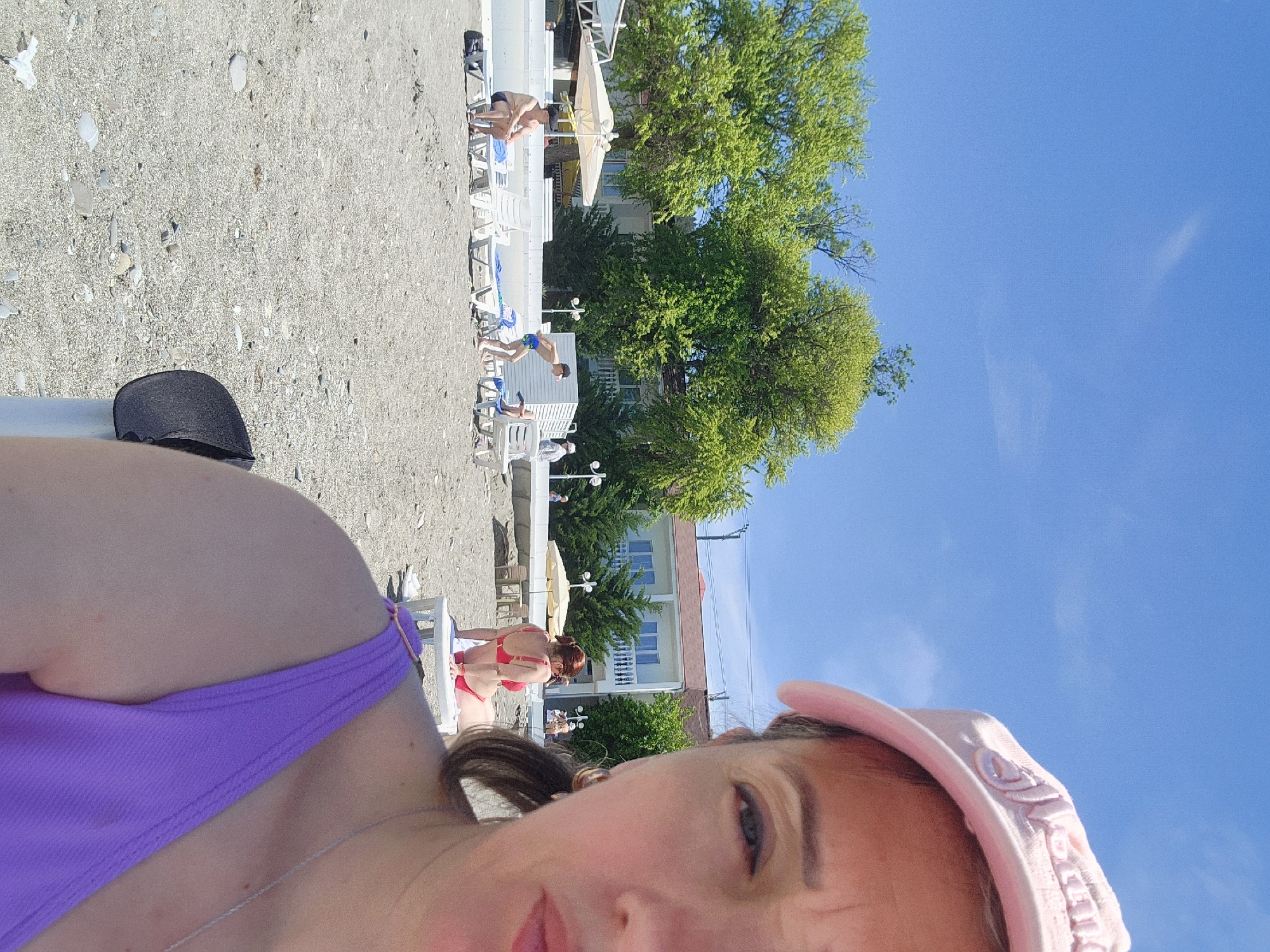 The image depicts a bright, sunny beach scene, captured as a selfie. The main subject, positioned in the foreground and appearing to take the photo, is a person wearing a purple swimsuit top and a pink baseball cap. Part of their face, including their eyes, nose, and mouth, is visible, along with their shoulder and a silver necklace. Their expression is neutral or slightly smiling. Indistinct embroidered text is visible on the pink cap.
In the mid-ground and background, a pebble and sand beach stretches out, populated with several white beach chairs or sun loungers. Other people are visible on the beach: one person in blue swim trunks is bending over near a chair, another in red swimwear is also near a chair, and a third in dark swim trunks is visible further back. A black object, possibly a hat, rests on the sand.
Behind the beach area, several lush green trees with dense foliage are prominent, indicating a vibrant natural environment. Beyond the trees, buildings, likely hotels or guesthouses, with balconies and windows, are visible. A beige sun umbrella is also present near the buildings. The sky above is a clear, bright blue, suggesting sunny weather during the day, likely midday or early afternoon given the strong lighting. The overall scene evokes a relaxed, vacation-like atmosphere. The original image is oriented 90 degrees clockwise.