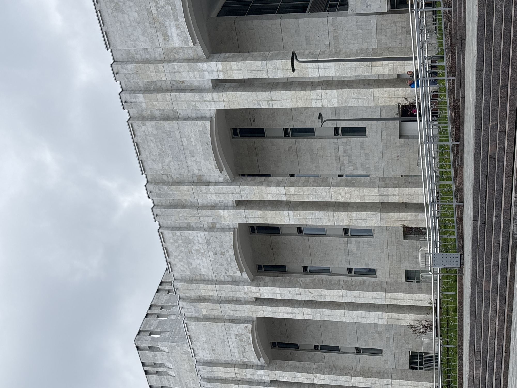 The image shows a building facade, likely in Perm, Russia, based on architectural style. The building is made of light-colored stone or concrete with a layered, tiered design, creating deep recesses for windows. Narrow, rectangular windows are visible within the recesses. A tall, metal lamppost stands in front of the building. A set of wide, granite steps run alongside the building, separated by a silver railing with grassy areas. A few people stand near the railing. The sky is overcast with grey clouds.