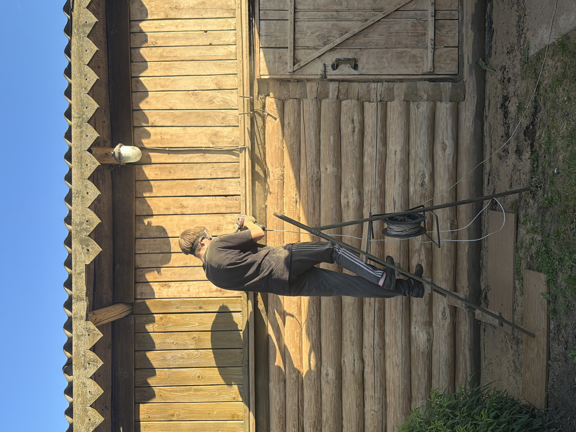 A young person wearing safety glasses and a dark t-shirt stands on a metal ladder, working on a log cabin. The person is holding a power tool, and dust is visible in the air around them, indicating they are sanding or cutting wood. The cabin is made of horizontal logs and has a wooden door on the right side. A spool of wire is attached to the ladder. The scene is lit by natural sunlight, casting long shadows. The location is outdoors, with grass and plants visible at the base of the cabin.