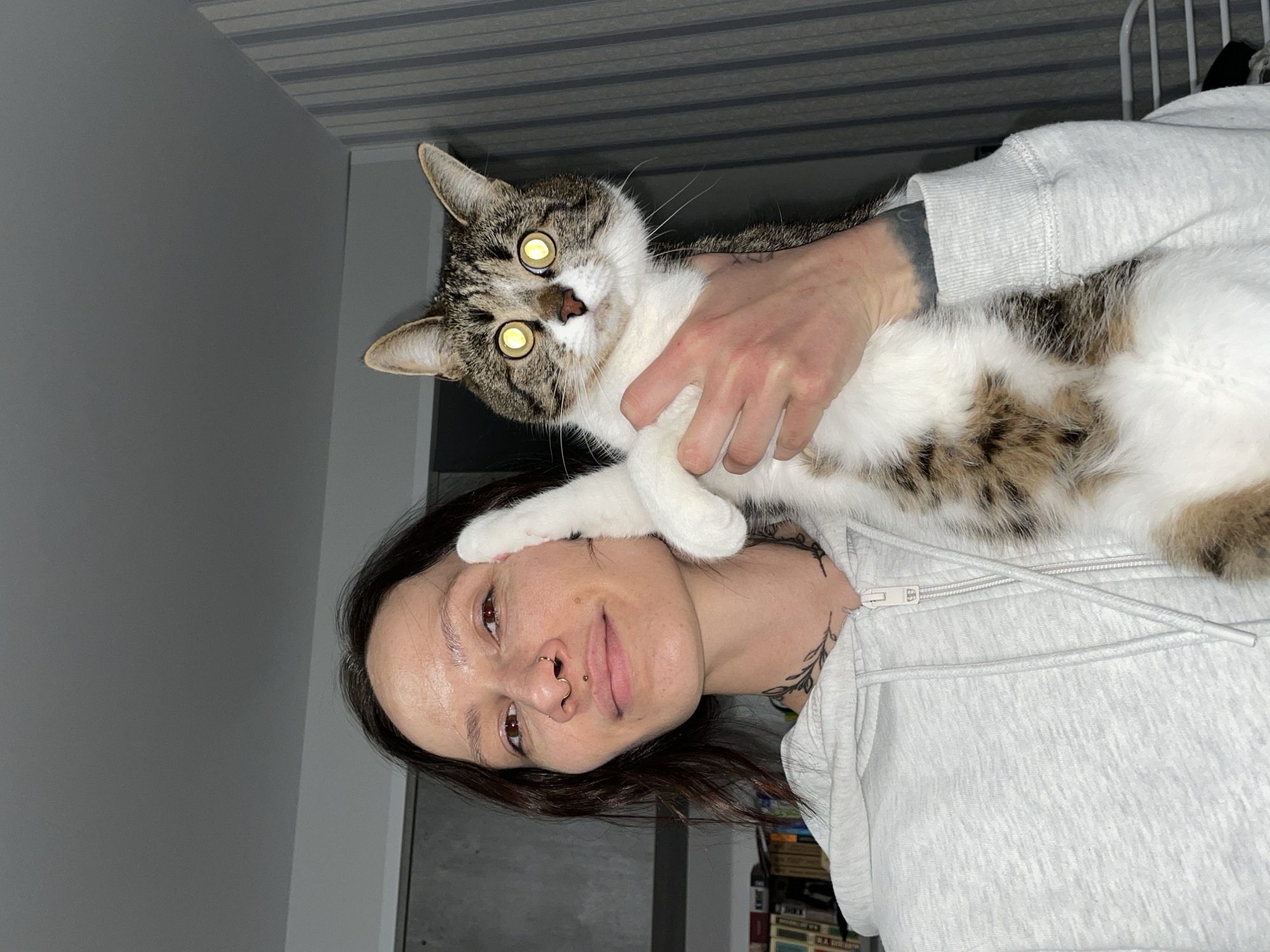 A woman is holding a cat, likely indoors. The woman has dark hair, light skin, and multiple piercings on her face, including a nose ring and a lip ring. She is wearing a light gray hooded sweatshirt. The cat is gray and white with glowing yellow eyes due to the camera flash. The woman is holding the cat close to her face. The background consists of a gray wall and ceiling, and a glimpse of a bookcase can be seen at the bottom of the frame. The setting is not Izhevsk, Russia.