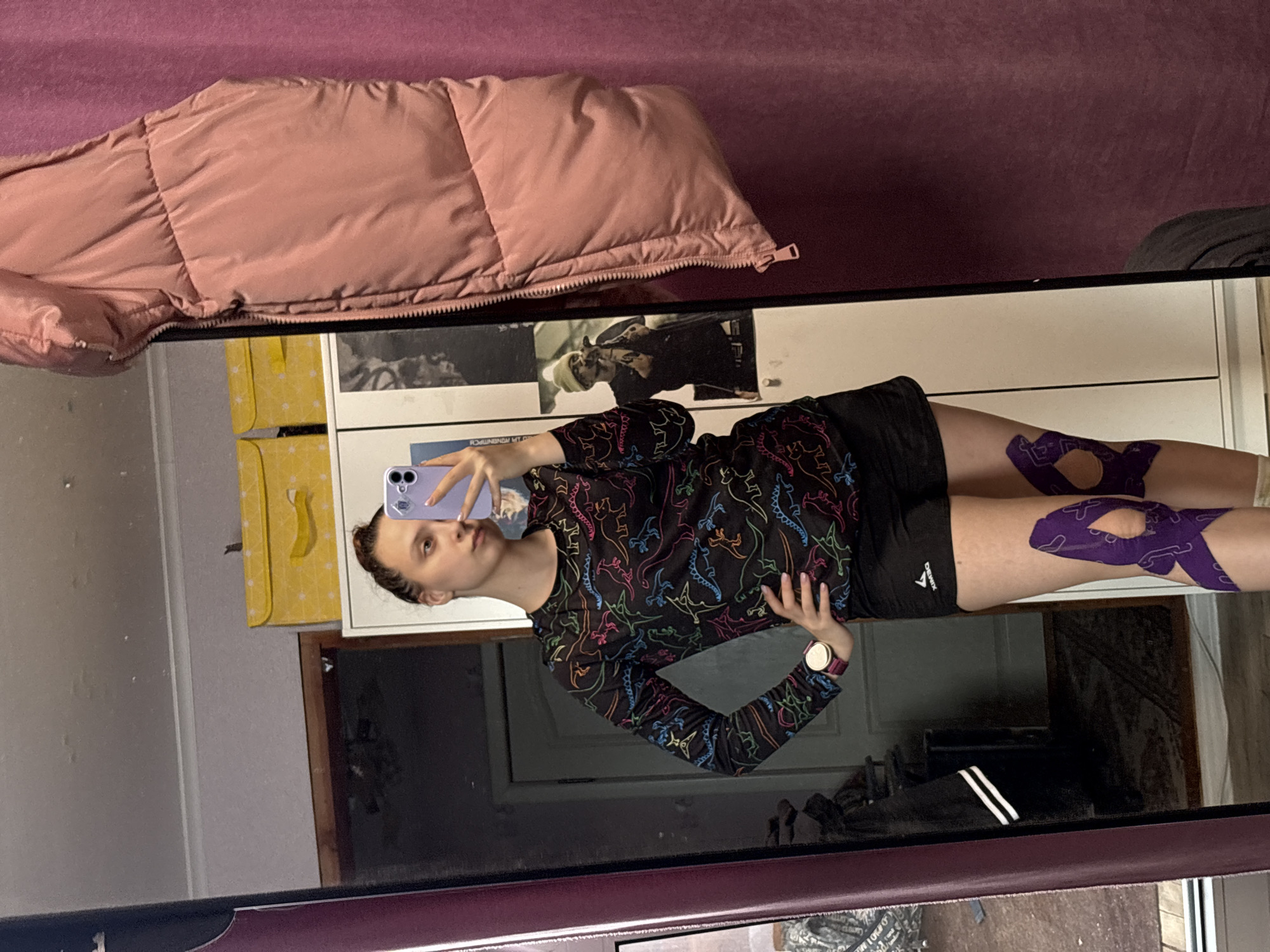 A young woman stands facing a mirror, holding a light purple smartphone to capture a selfie of her reflection. She has light brown hair pulled back and a neutral expression as she looks at the phone. She is dressed in a black long-sleeved shirt adorned with colorful outlines of dinosaurs, and black athletic shorts with "DEMIX" visible on the left leg. A smartwatch with a pink band and gold face is worn on her left wrist.
Notably, both of her thighs are wrapped with purple kinesiology tape around the knee area, suggesting athletic activity, injury support, or muscle recovery. The background, visible in the mirror's reflection, includes a large pink puffer jacket, white furniture (likely a dresser or shelf unit), and yellow decorative bags or boxes. A poster or picture depicting a person in a beanie is also visible on the furniture. The setting appears to be an indoor, personal space such as a bedroom or dressing area.