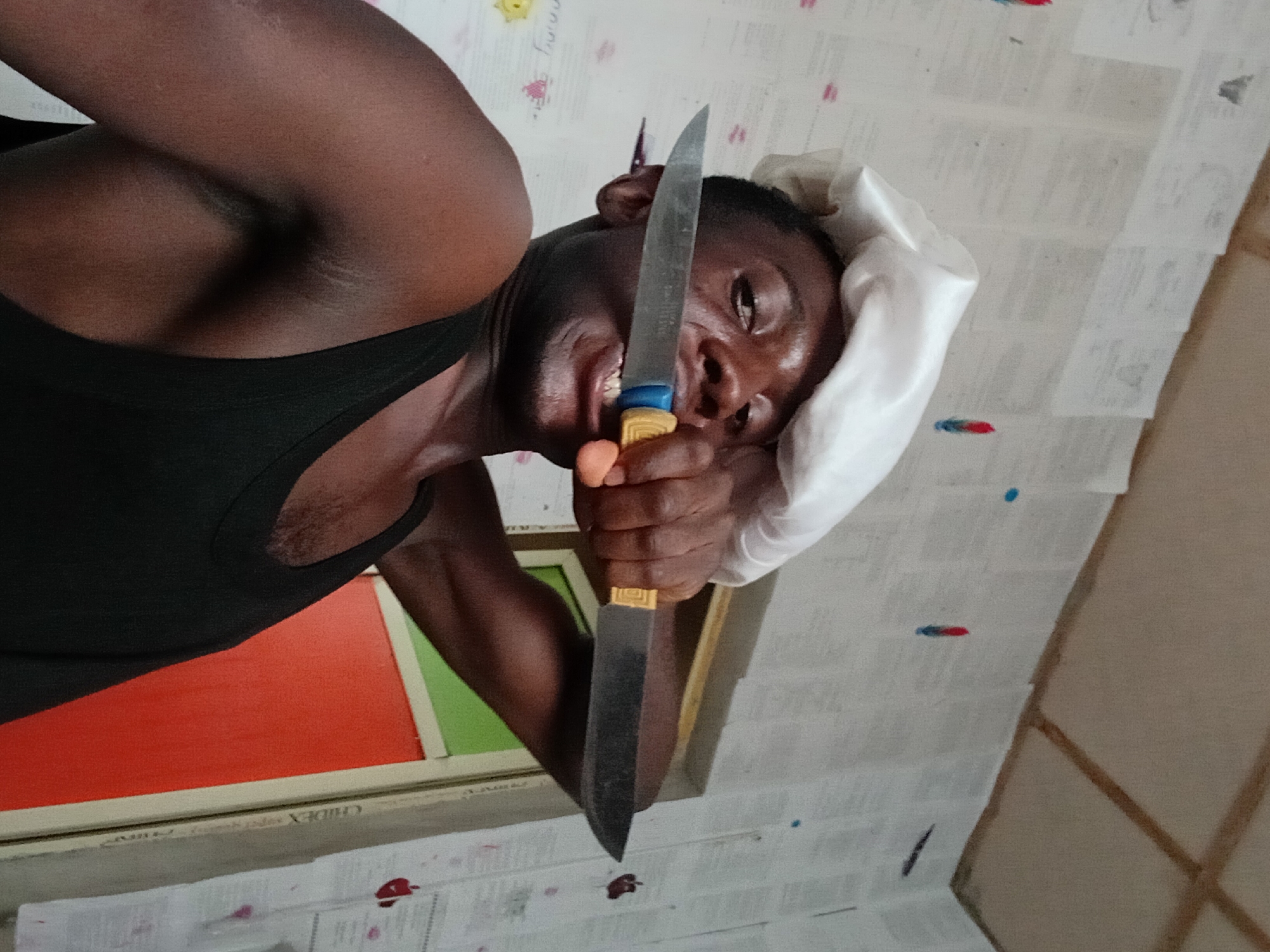 A dark-skinned man, wearing a white chef's hat and a black tank top, holds a large kitchen knife with a blue and yellow handle in his right hand, positioning the blade vertically in front of his face. The man is smiling broadly, exposing his teeth. He is indoors in what appears to be a room with walls covered in paper. In the background, a framed picture with orange and green segments is partially visible. The scene is possibly in Akwete, Nigeria.