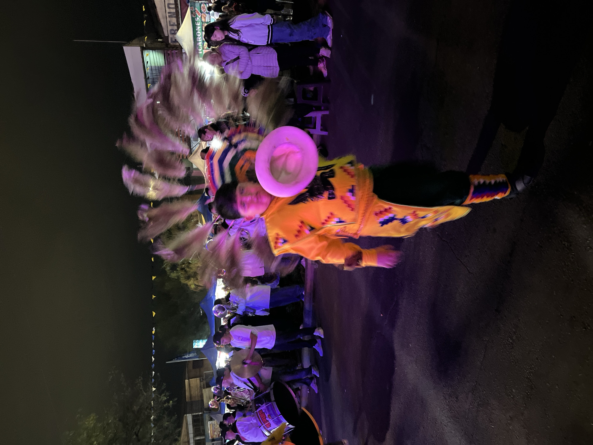 This is a photograph taken at night, likely at an outdoor event or festival in Arica, Chile. In the foreground, a young person is captured in motion, wearing a vibrant yellow costume adorned with colorful geometric patterns. They are holding a pink, circular object, possibly a hat or prop, in their right hand, which is blurred by movement. The costume has a headdress made of long, feathery plumes that extend upwards and to the right, also blurred by the motion of the performer. The person is wearing dark pants and colorful leg coverings with similar geometric designs.
In the middle ground and background, a crowd of people are visible, observing the performance. Some are holding instruments, including drums and what appear to be brass instruments, suggesting a musical procession or performance. Tents and structures, possibly market stalls or stages, are present in the background, illuminated by artificial lighting. The ground is paved and dark, with reflections visible from the lighting. The overall scene is dynamic and festive, with a sense of movement and community gathering. The low-light conditions and the motion blur indicate the photograph was taken with a relatively long exposure time, characteristic of nighttime photography at events.