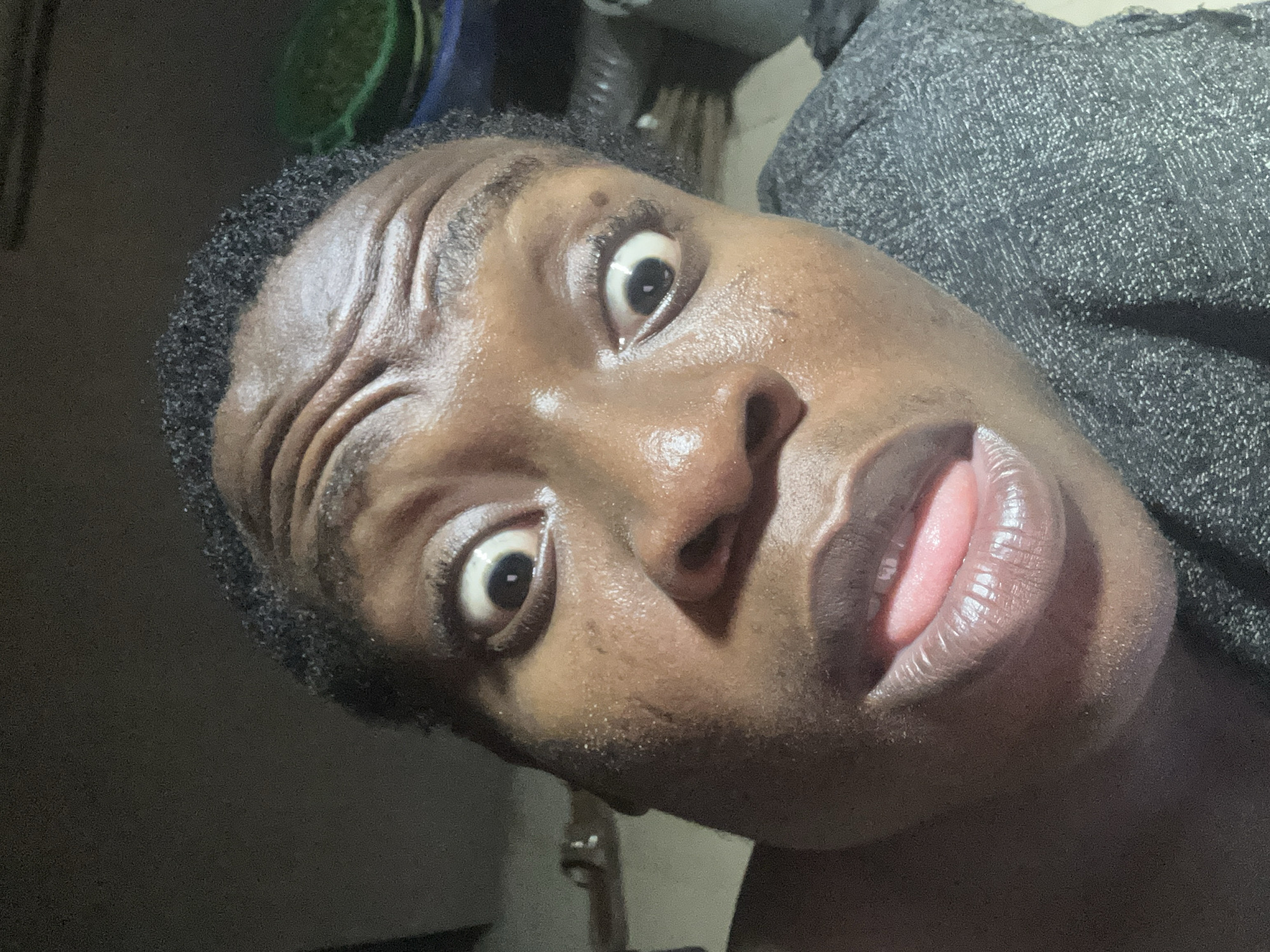 A close-up, distorted-angle photograph shows a Black person with their head tilted slightly. The person has short, dark hair and dark skin that appears slightly glossy. Their eyes are wide open and looking upwards, with the left eyebrow raised. Their mouth is slightly open, revealing a portion of their tongue. The background is a mix of dark and light areas, with some blurred shapes that appear to be objects in a room. A gray, textured fabric can be seen on the right side of the frame. The location is Unknown, Unknown.