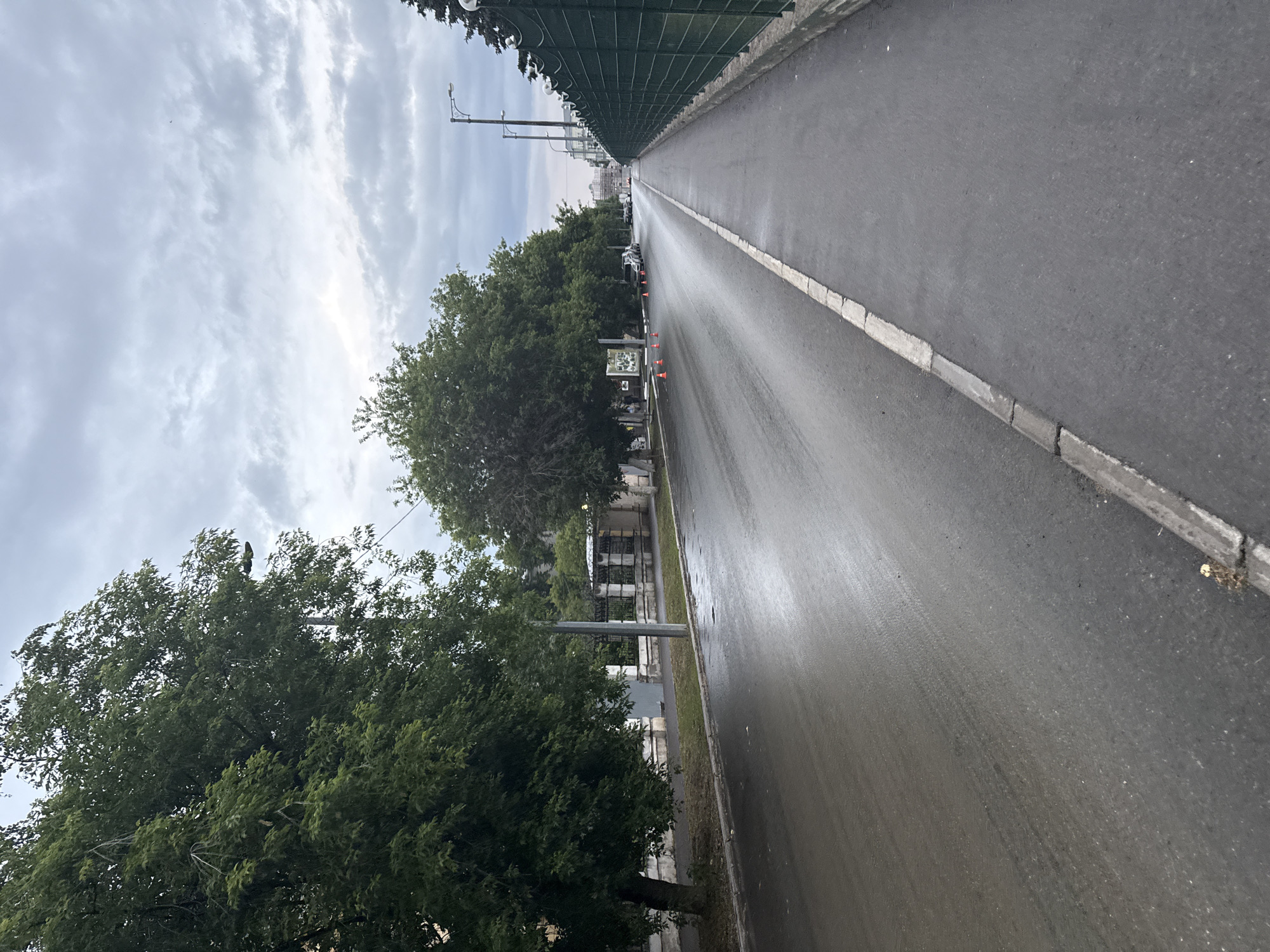 The image depicts a wet asphalt road in Kazan, Russia, under a cloudy sky. On the left side of the road, a row of lush green trees lines the sidewalk, which is separated from the road by a concrete curb. Further back, a decorative fence and buildings are visible. Several orange traffic cones are placed on the wet road, suggesting a potential hazard or a temporary closure. A few cars are parked along the side of the road in the distance. On the right side of the image, a paved sidewalk runs alongside a dark green metal fence, also bordered by a concrete curb. The overall scene suggests a post-rain environment in an urban setting.
