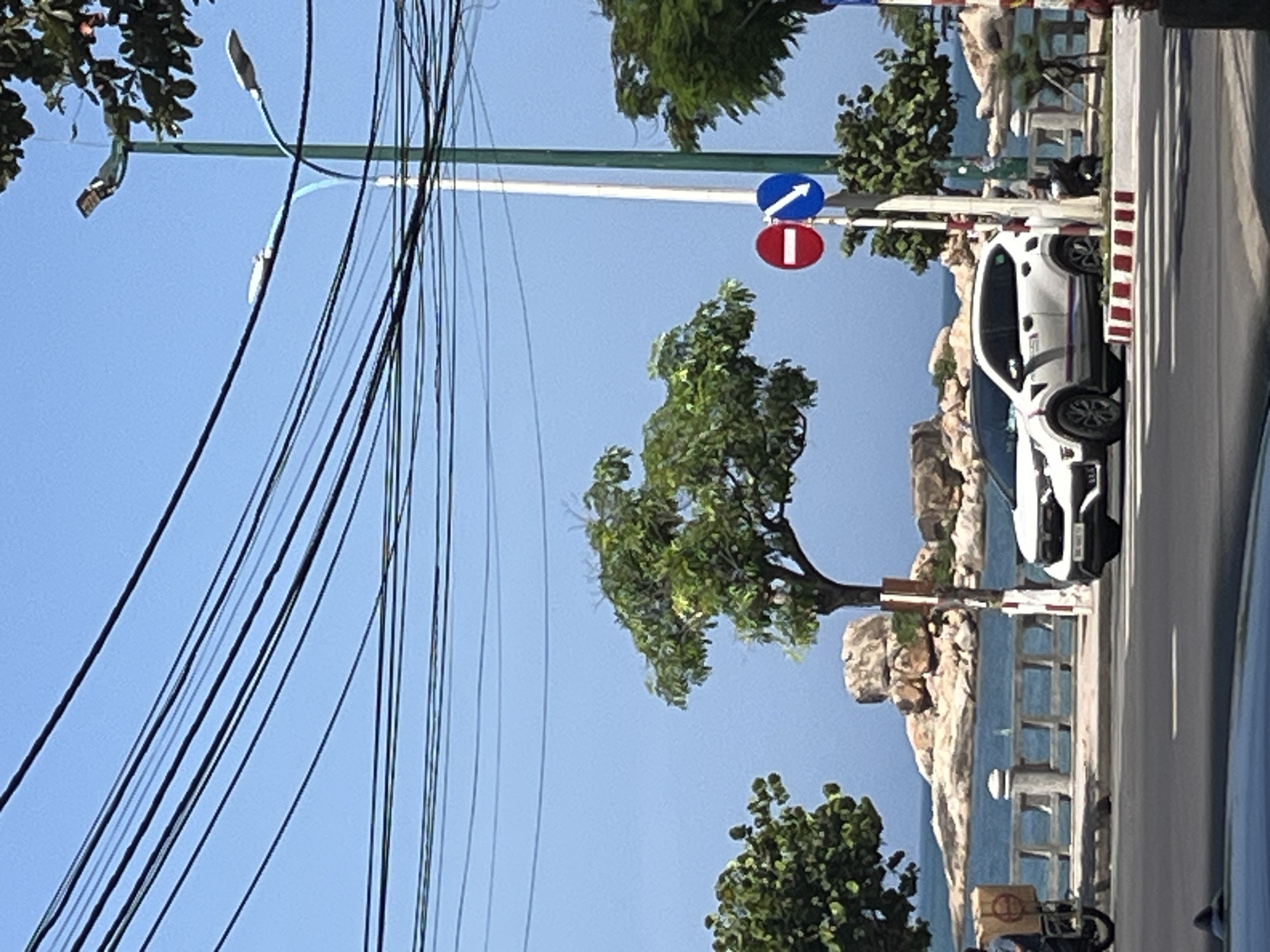 The image depicts a bright daylight scene on a coastal road in Bac Nha Trang Ward, Vietnam. The upper portion of the frame is dominated by a clear, light blue sky crisscrossed by multiple black utility lines. Two street lighting poles are visible: a tall green pole on the right and a white pole with a light blue curved arm holding two light fixtures in the center.
In the midground, beyond the road and a concrete barrier, a blue ocean extends to the horizon. Several large, light brown rocky formations are scattered across the water, resembling small islets or coastal outcrops. Green-leafed trees are interspersed throughout the scene, with a prominent tree positioned centrally and other foliage visible on both sides of the road.
Two circular traffic signs are mounted on a white pole near the center-right: a red "Do Not Enter" sign with a white horizontal bar, positioned above a blue mandatory sign with a white arrow pointing diagonally downward to the right.
The foreground consists of a dark asphalt road featuring a white crosswalk. The curb bordering the road is painted with alternating red and white stripes. A white crossover SUV, possibly a Honda HR-V, is stopped or parked on the right side of the road, facing left, with blue and red stripe decals on its lower body panels. On the far left, the rear of a motorcycle with a large brown delivery box, featuring a circular reddish logo, is partially visible, with a person seated on it. Another vehicle's dark roof is visible at the very bottom of the frame. An individual is seated on the ground or curb on the right side of the road, partially obscured by the white SUV and a tree.
