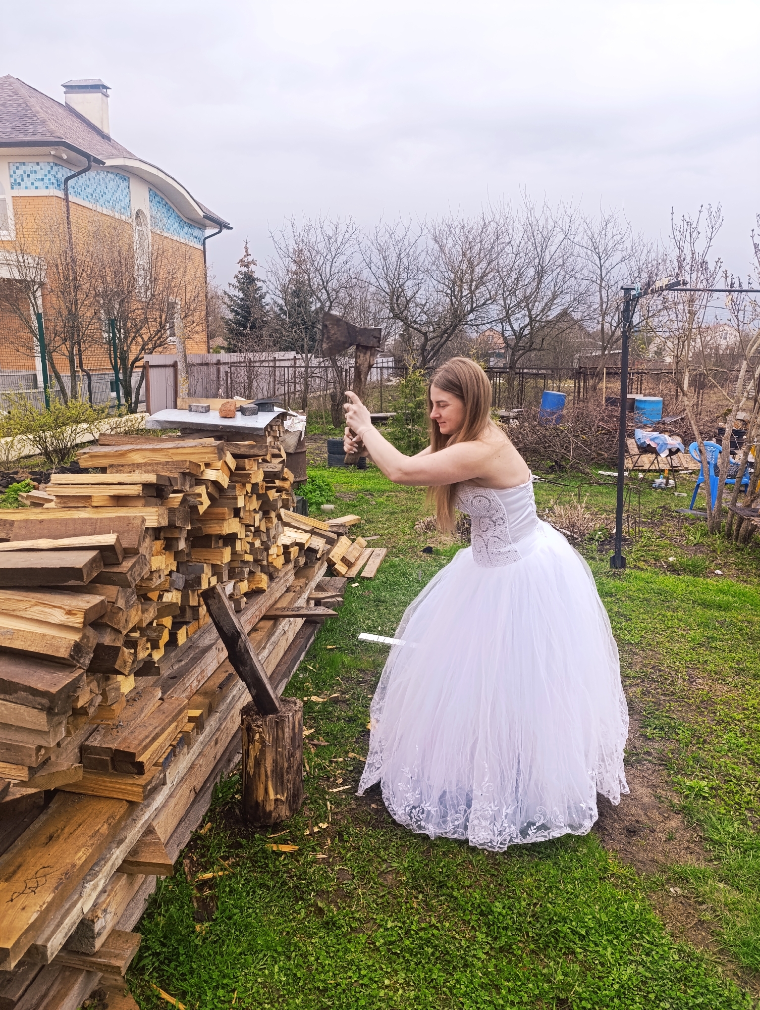 A woman in a white wedding dress is splitting wood with an ax in a backyard in the city of Unknown, Unknown. A large pile of split wood sits next to her, with a stump acting as a chopping block. The backyard is grassy and includes trees, a fence, and a house.