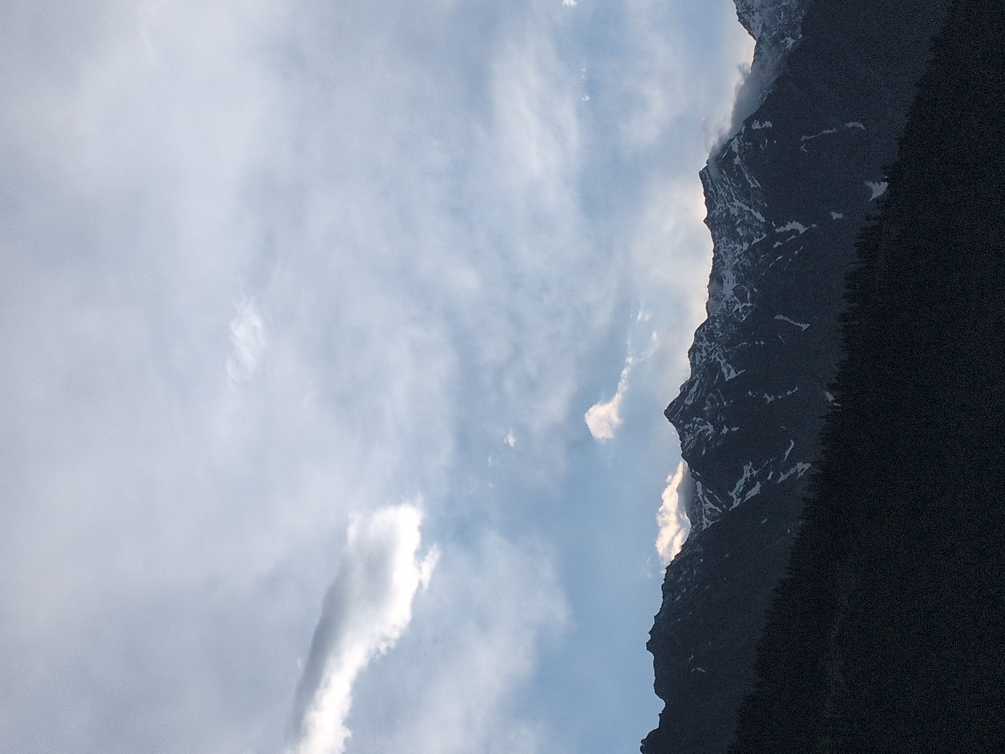 The image shows a panoramic view of snow-capped mountains under a cloudy sky. The mountains are dark and jagged, with patches of white snow on their peaks and slopes. A dense forest of dark trees covers the lower part of the mountains, creating a stark contrast with the lighter sky. The sky is filled with various shades of gray and white clouds, with some patches of blue sky visible in the distance. The lighting suggests it could be early morning or late evening, with the sun low on the horizon, casting a diffused light on the scene. The image appears to be taken from a vantage point looking up at the mountains. There are no people or animals visible in the image. The overall atmosphere is one of natural grandeur and solitude. The location is Vashisht, India.
