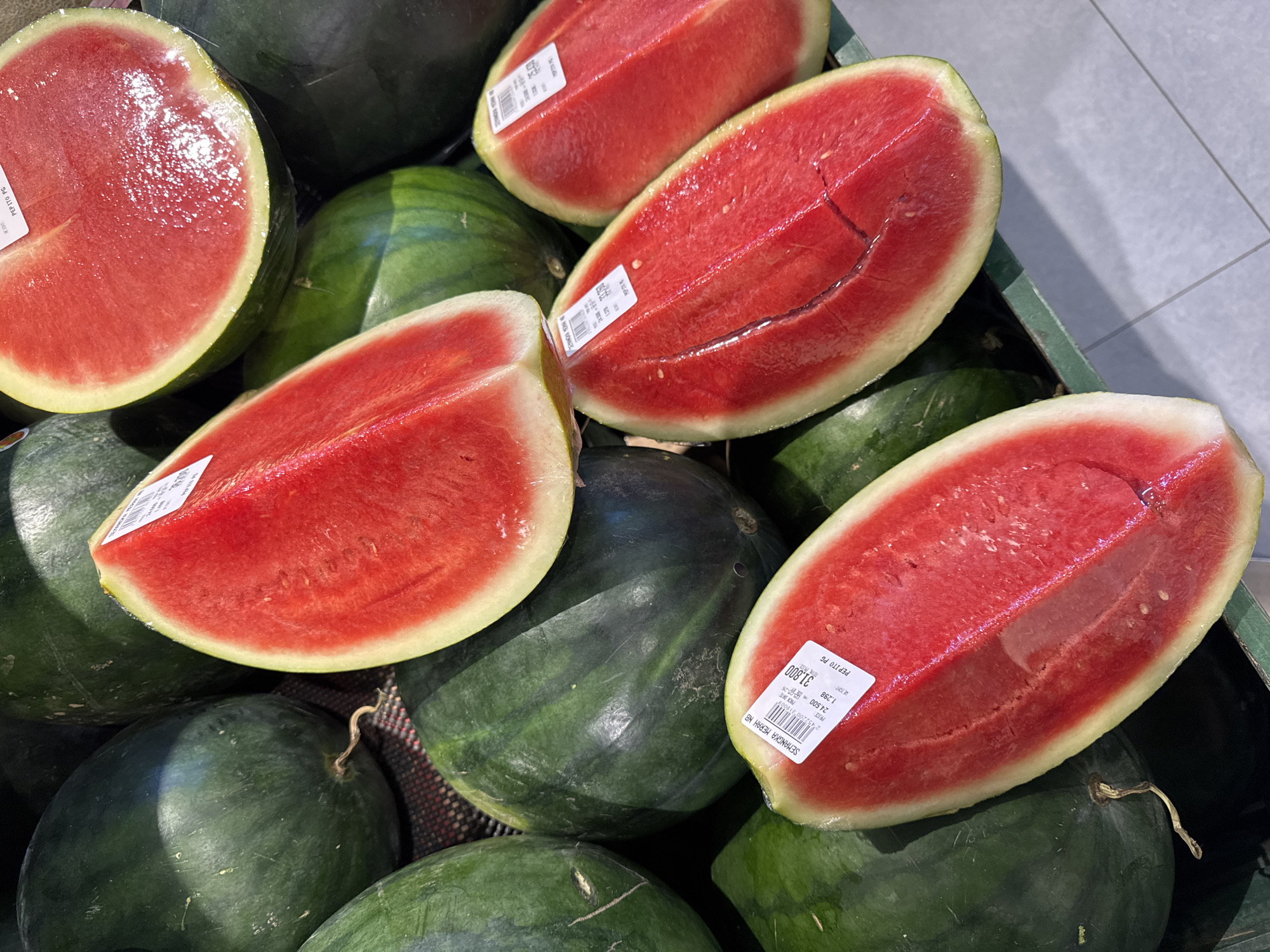 A close-up shot shows a display of watermelons at a market, likely located in Kuta Selatan, Indonesia, given the Indonesian text on the price labels. A mix of whole watermelons and cut portions are piled together in a green crate. The whole watermelons have a dark green rind, while the cut portions reveal a bright red flesh with visible seeds. Price labels with both numerical and Indonesian text are attached to several watermelons. The lighting is bright, emphasizing the vibrant colors and textures of the fruit.