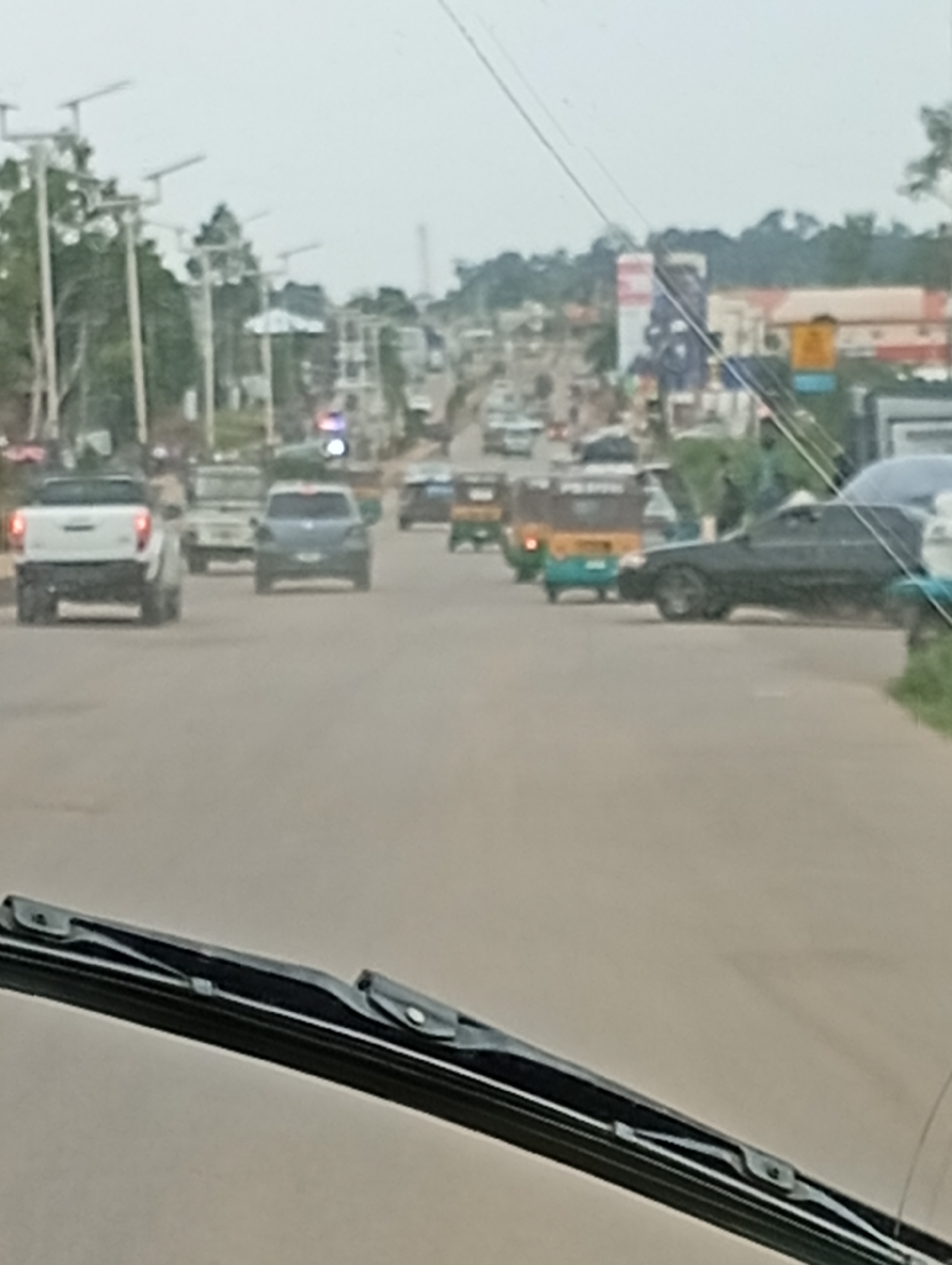 The image shows a street scene in Bukuru, Nigeria, likely a commercial or residential area given the presence of buildings, power lines, and various vehicles. The perspective is from inside a vehicle, with the windshield wipers visible in the foreground. The road appears to be paved, with multiple lanes of traffic. Several vehicles are present, including a white pickup truck, a dark sedan, a light-colored SUV, and several auto-rickshaws, also known as Keke Napep. Some of these vehicles are in motion, while others appear to be stationary. The background reveals a mix of buildings, some with colorful signage, and lush green vegetation, suggesting a tropical or subtropical climate. Power lines and poles line the street, common in urban infrastructure. The overall impression is that of a busy street in a developing urban environment.