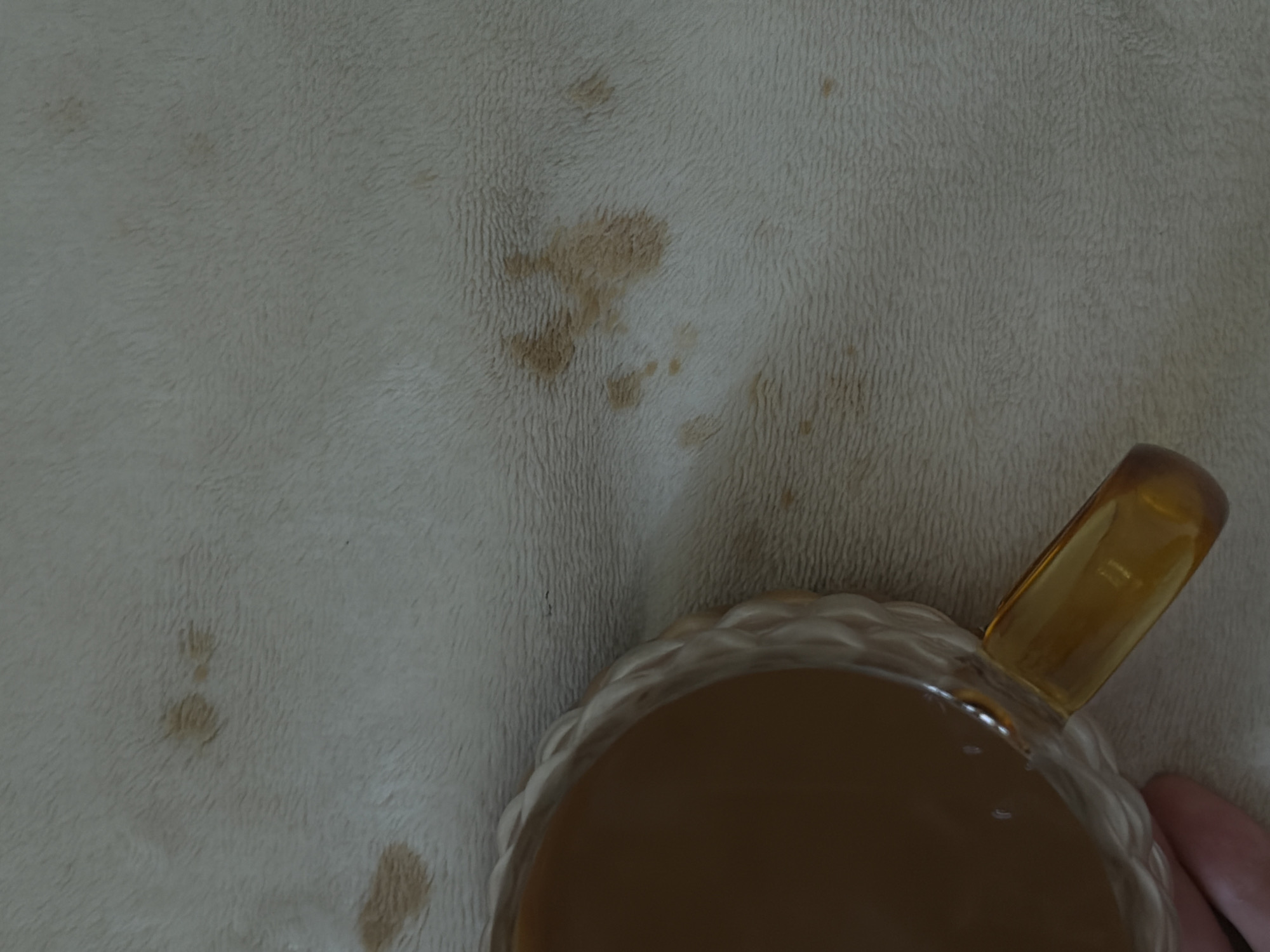 The image shows a beige, soft, textured surface with several brown stains scattered across it. A portion of a glass mug is visible in the lower right corner, containing a light brown liquid. A human finger is partially visible near the mug. It is not possible to determine the location in Izhevsk, Russia, or any further details about the environment or interactions from this image.