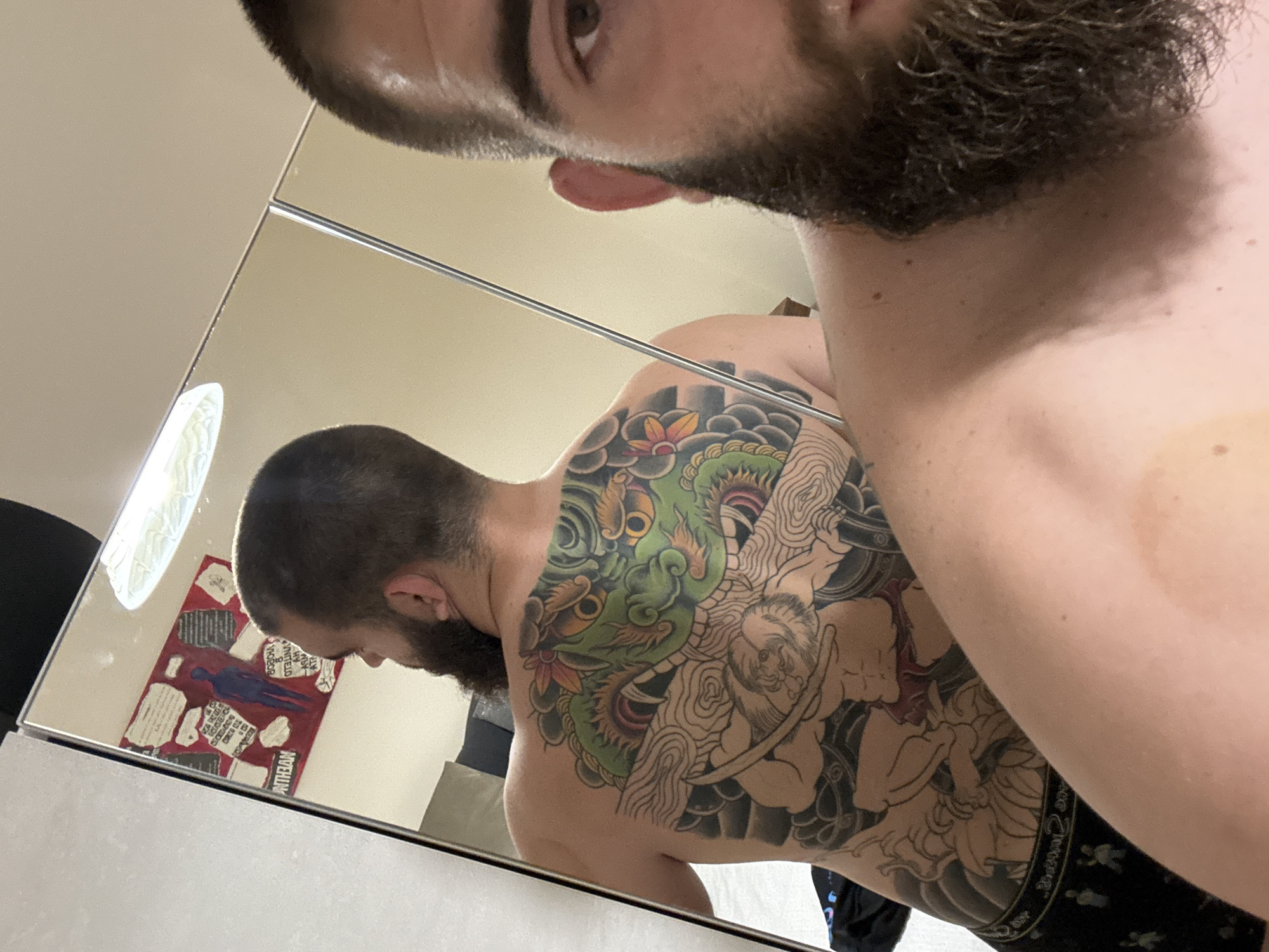 A man with a beard is taking a selfie in a mirror, partially showing his face and upper body. The reflection reveals his bare back and side, which feature a large, colorful tattoo. The tattoo includes a green dragon-like creature, flowers, and other design elements. The room appears to be a bathroom, with a poster visible in the mirror's reflection. There is no indication of the location in the city of Stara Zagora, Bulgaria.