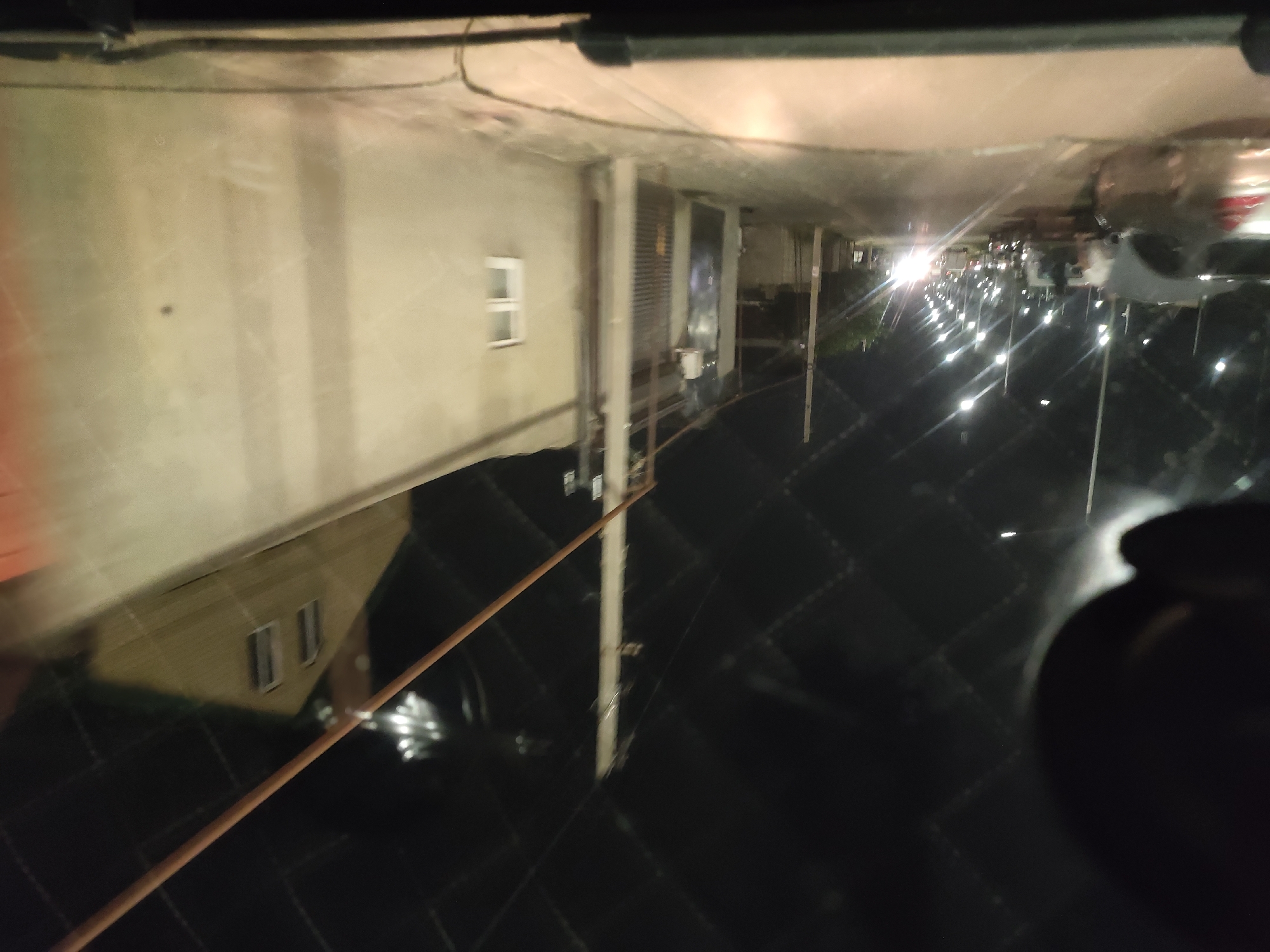 This image captures a street view in Leninkent, Russia, at night. The scene is dominated by a road lined with streetlights that illuminate the asphalt and the surrounding environment. Multiple vehicles are present on the road, some parked and others in motion. On the right side of the frame, a light-colored wall runs along the street, with a small white-framed window visible on it. Beyond the wall, a sloped roof of a building can be seen, with two dark-paned windows. The overall impression is that of an urban street scene at night, with artificial lighting playing a significant role in defining the visibility and mood. The sky is dark, with a patterned ceiling overhead, suggesting the scene might be within an enclosed or covered area, or perhaps a reflection.