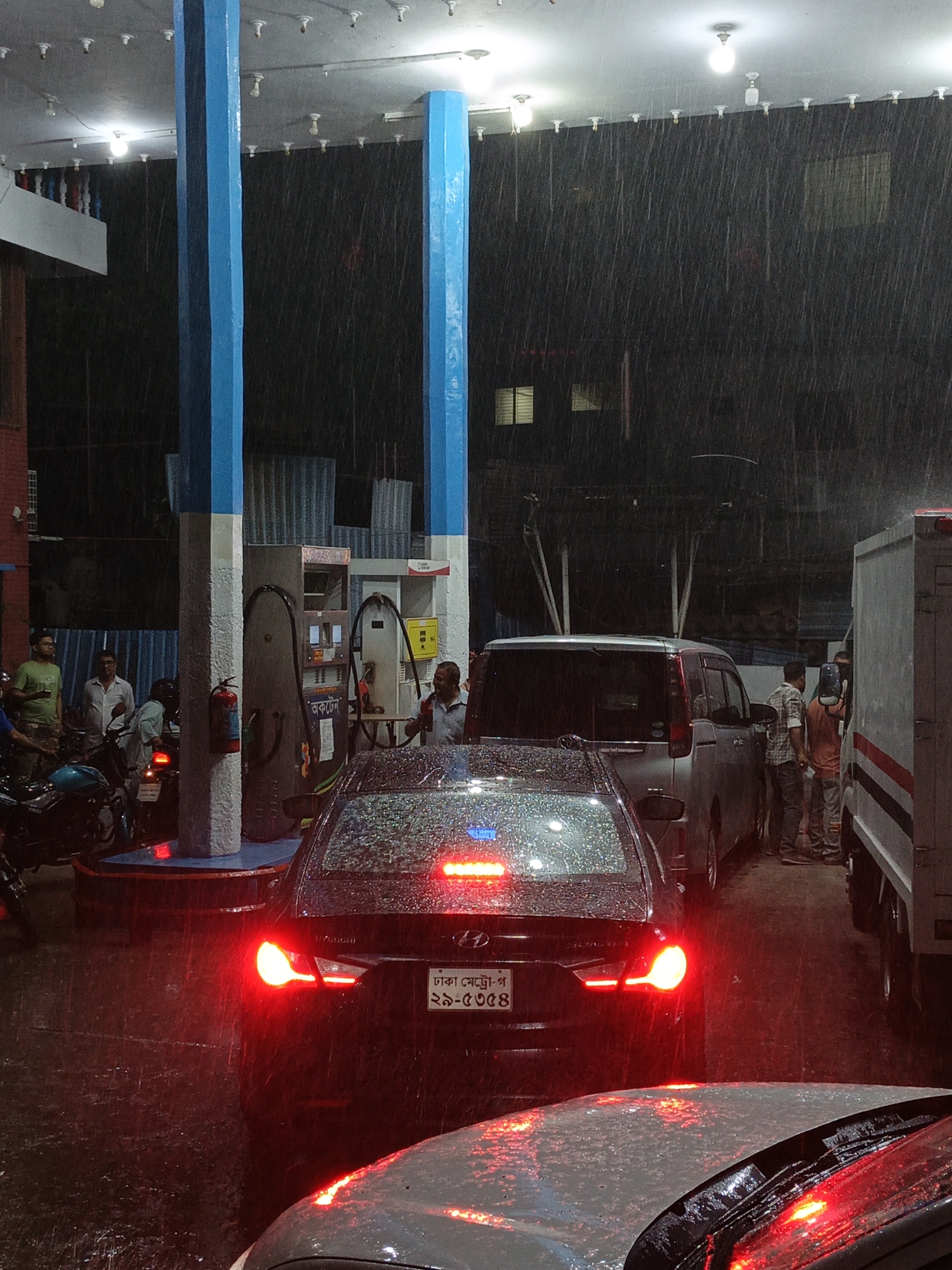 At a gas station in Dhaka, Bangladesh, it is raining heavily at night. Cars and motorcycles are lined up at the pumps. Several people are visible: gas station attendants, drivers, and passengers. Streetlights and the gas station's overhead lights illuminate the scene. One black sedan in the queue has its brake lights illuminated. The car in front of it is also visible, with water droplets covering the roof. A delivery truck is on the far right of the frame. Blue and white painted columns support the gas station awning.