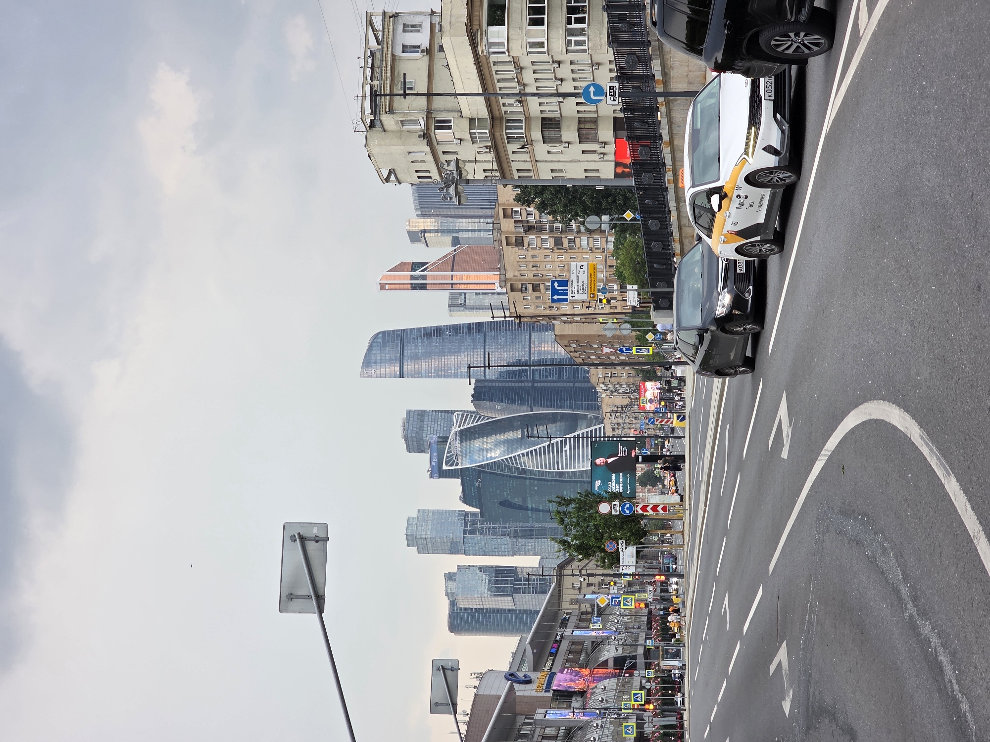 This is a street-level, medium-shot, eye-level, horizontal photograph of a busy road in Moscow, Russia, with the Moscow City skyscrapers in the background. The sky is overcast with gray clouds. The foreground shows a multi-lane asphalt road with white lane markings. Several cars are on the road, including a white Yandex Taxi with a yellow and orange livery, and two dark-colored sedans. In the middle ground, there are buildings of varying architectural styles, including modern apartment blocks and older structures. Various street signs and traffic lights are visible, along with a pedestrian crossing. The background is dominated by the iconic Moscow City International Business Center, featuring several distinct skyscrapers with modern glass facades, including the distinctive twisted "Eurasia" and "Naberezhnaya Tower". The overall impression is of a bustling urban environment in Moscow.