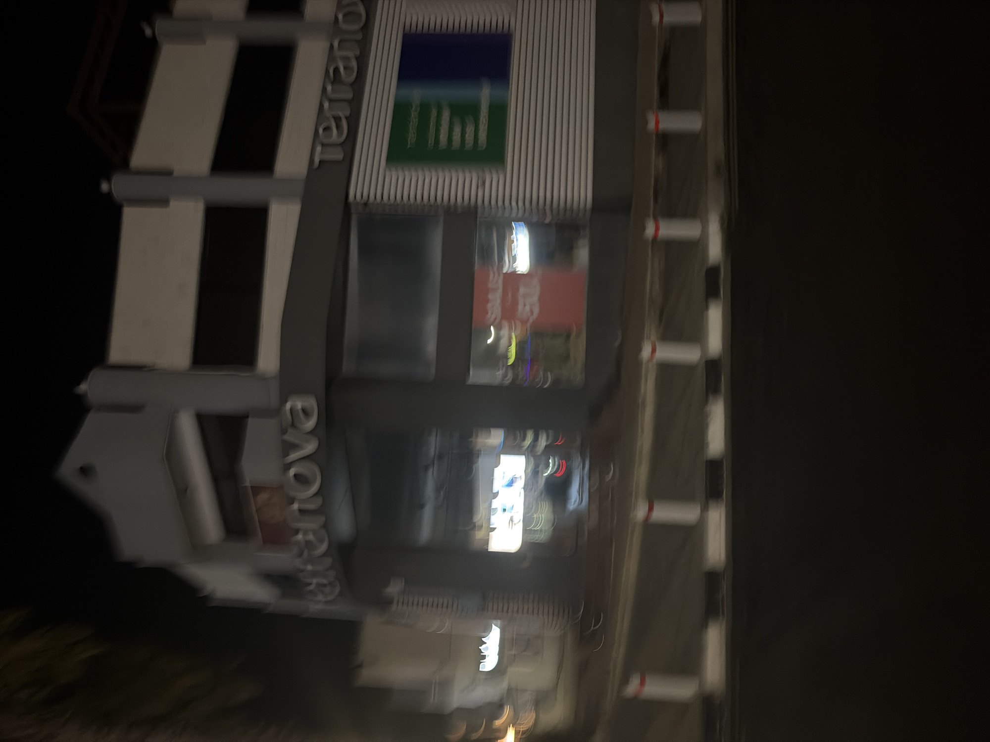 The image shows a blurred night view of the Terranova store facade in Agios Athanasios Municipality, Cyprus. The store features a prominent sign with the word "Terranova" in large, silver lettering. The building has a modern architectural design with horizontal and vertical elements. A green and blue sign is visible on the right side of the facade, possibly displaying store information or promotions. In the foreground, a road is visible with white bollards and black and white markings. The overall scene is dimly lit, suggesting it was taken at night.