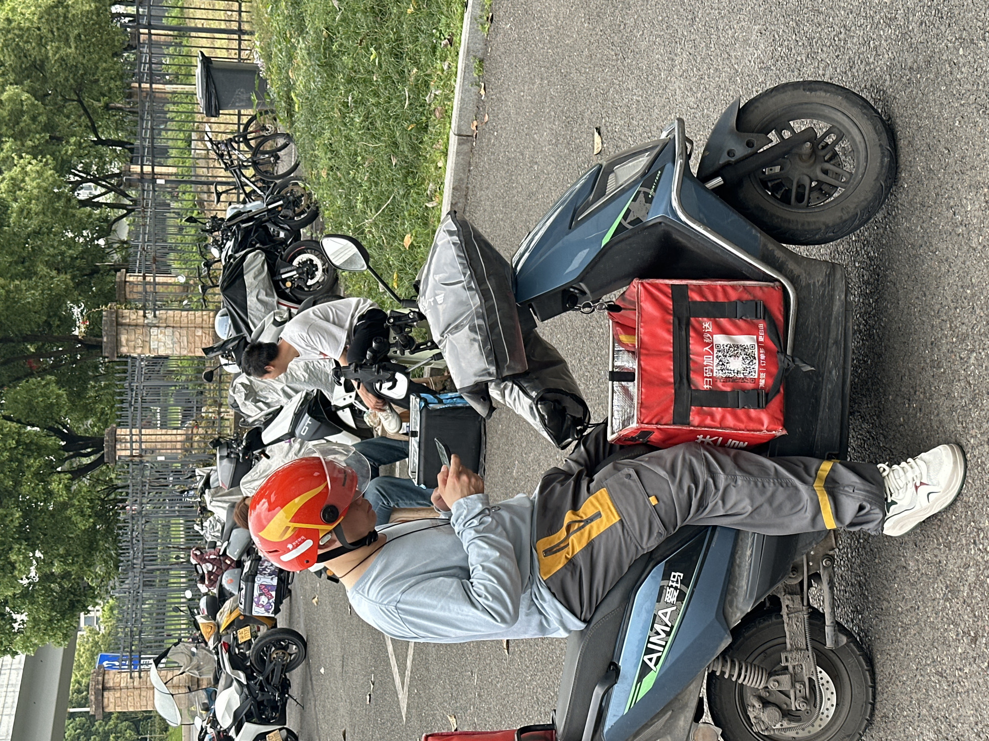 The image depicts a group of delivery drivers resting on their scooters in Jiangjin, China. A delivery driver in the foreground, wearing a red helmet with yellow stripes, a light blue long-sleeved shirt, and gray pants with yellow accents, is seated on a blue AIMA scooter, looking at a phone. A red delivery box is attached to the back of the scooter, displaying a QR code. Several other scooters, some with covers, are parked behind, along with other delivery drivers. A black metal fence and some trees are visible in the background. The scene is situated on an asphalt surface, likely a parking area.