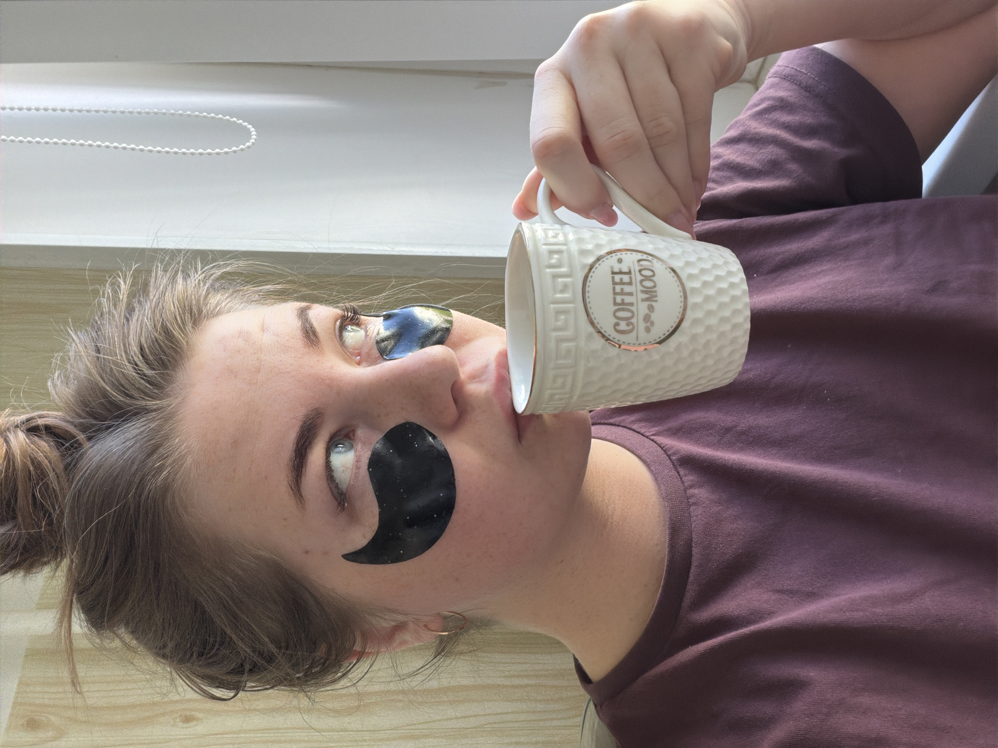A fair-skinned woman with brown hair pulled back in a messy bun is holding a white mug that reads "COFFEE MOOD" in her right hand. She has black eye masks under each eye. She is wearing a maroon t-shirt. The photo is taken indoors, with a window and white blinds in the background. There is no indication of the city of Chaikovsky, Russia.