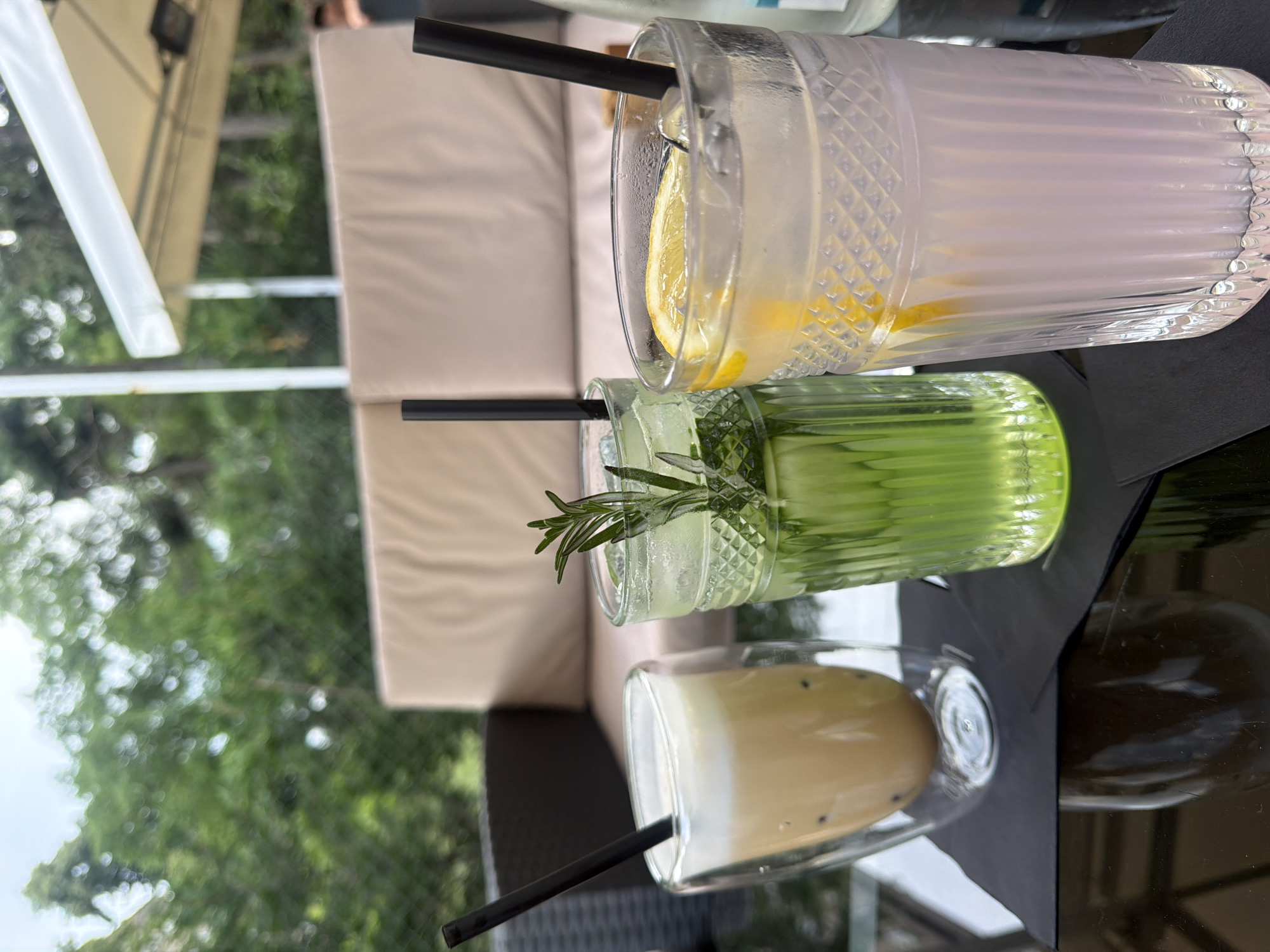 Three distinct beverages are presented on a dark, reflective glass table in an outdoor setting. The leftmost beverage is contained within a clear, double-walled glass, displaying a light brown, opaque liquid topped with white foam, and dark, spherical elements are visible at the bottom. A black straw is inserted. The central beverage is in a clear, faceted glass with a diamond-cut pattern at the top and vertical fluting; it holds a vibrant green, translucent liquid with ice cubes and is garnished with a sprig of rosemary and what appears to be cucumber slices. A black straw is also present. The rightmost beverage, in a matching faceted glass, contains a pale pink/purple, translucent liquid with ice cubes and is garnished with a yellow lemon slice, also with a black straw. Two black square napkins are placed under the glasses.
In the midground, a dark brown, possibly rattan or wicker, outdoor sofa with light beige cushions is visible. Further back, additional light beige seating is present. The background consists of out-of-focus, dense green foliage, indicative of a natural, park-like environment. A segment of a white tent or awning structure with white support poles is visible on the upper right, partially obscuring a blurry, indistinct human figure (head and shoulder) on the far right. Subtle mesh-like fencing or netting is discernible within the blurred green background. The scene depicts an outdoor cafe or bar situated within a verdant, park-like area in Sofia, Bulgaria. Specific architectural or landmark features for precise venue identification are not discernible within the image.