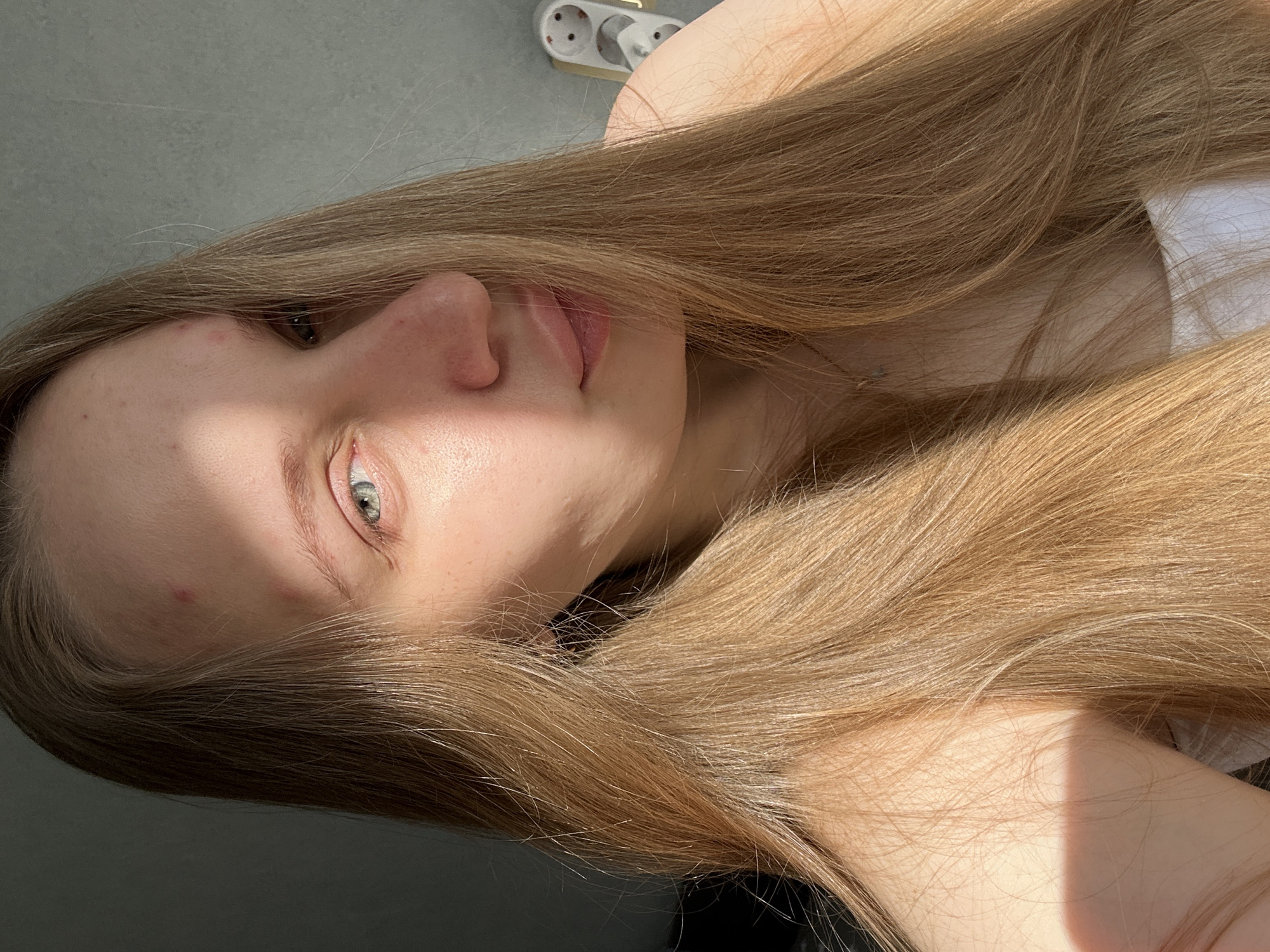 This close-up portrait, oriented correctly, features a young person, likely female, indoors. The subject's face is partially visible, primarily the left side, including a blue-grey eye, nose, and mouth, looking directly at the camera. Long, straight, light brown to blonde hair flows around the face and upper body, covering much of the frame.
The scene is lit by strong, natural daylight, creating pronounced shadows and highlights. A distinct shadow falls across the upper part of the face, contrasting with the brightly lit eye and nose. The skin shows natural texture and a few minor blemishes on the forehead and nose. The expression is neutral and direct.
In the background, a light grey wall or surface is visible, along with a white European-style electrical outlet featuring two circular sockets and a grounding pin, indicative of an indoor setting. A darker grey/black surface is present in the lower part of the frame. The strong light suggests a sunny day, likely morning or afternoon. The electrical outlet is consistent with those found in Russia, fitting the location context of Chaikovsky. There is no visible text.