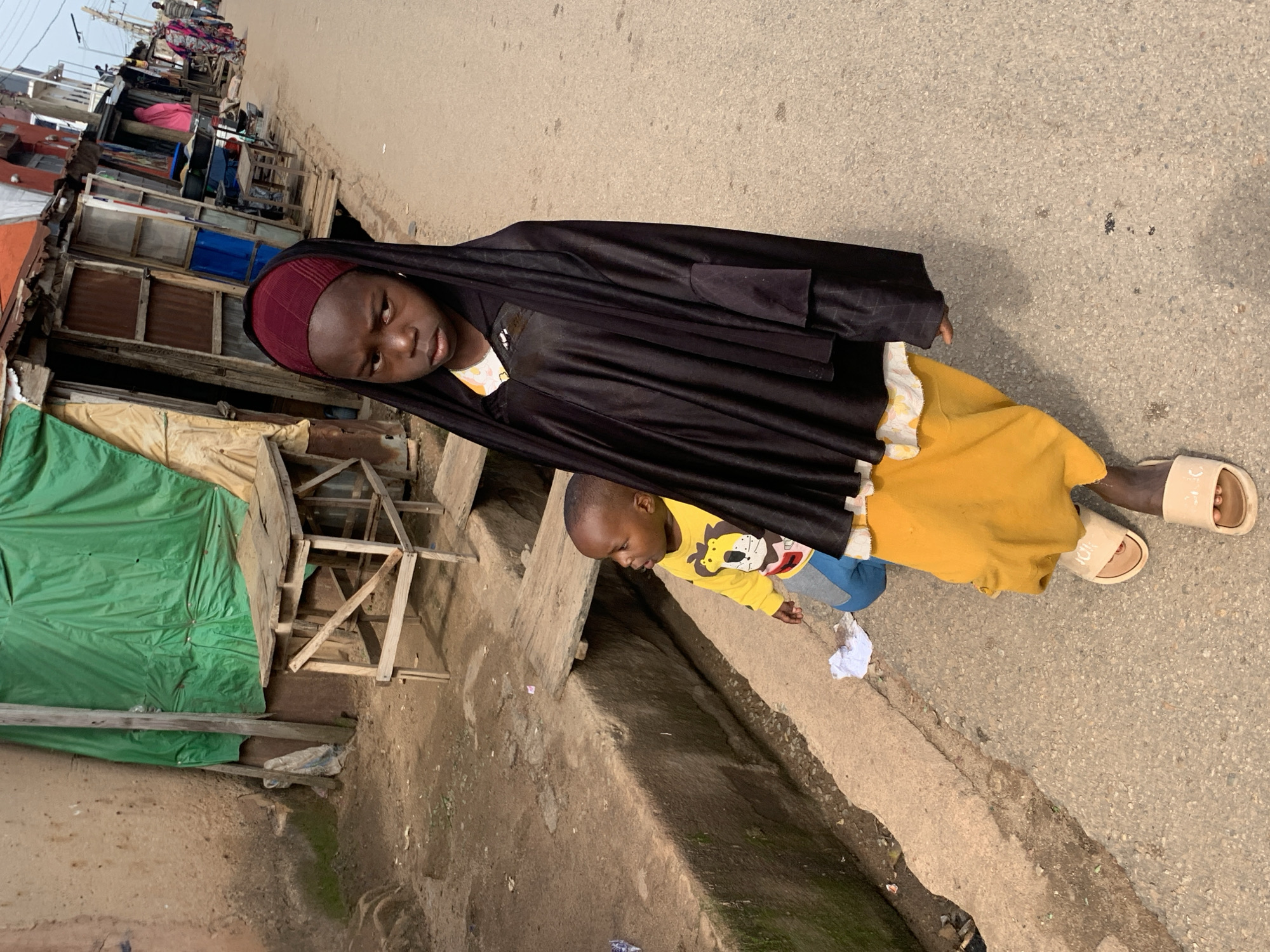 In Jos, Nigeria, a young girl, possibly around 8-10 years old, stands on a paved street, her expression stern. She wears a dark headscarf and a loose-fitting black garment over a yellow skirt. Her exposed feet are clad in beige slide sandals. Behind her, a toddler in a yellow long-sleeved shirt with a cartoon character and gray pants looks to the side with curiosity. The toddler is partially obscured by the girl's clothing.
The environment suggests a market or street-side vendor area. To the left of the street, there is a raised, rough-textured concrete walkway or drainage channel. Beyond this, a wall of earth and rough stone is visible, with patches of moss. A rudimentary wooden structure, possibly a stall or table, is positioned against this wall, covered by a green tarp. Further down the street, a row of small shops with corrugated metal roofs and wooden fronts are visible. Some stalls are open, displaying various items. In the background, other people are walking along the street, and some buildings are visible. The overall impression is of a bustling urban street scene.