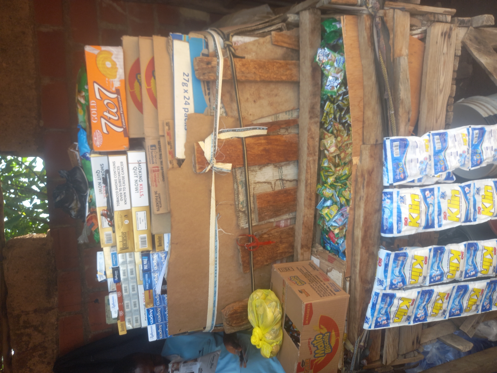 The image shows a retail stall constructed from wood and cardboard, displaying various goods. The stall is located outdoors, with a brick wall and foliage visible in the background. The items for sale include packs of cigarettes, boxes of biscuits labeled "Malto Biscuits", and multiple packets of "Klin" detergent hanging from the stall. A person, partially visible on the left, is wearing a light blue shirt. The overall scene depicts a small, informal market setting.