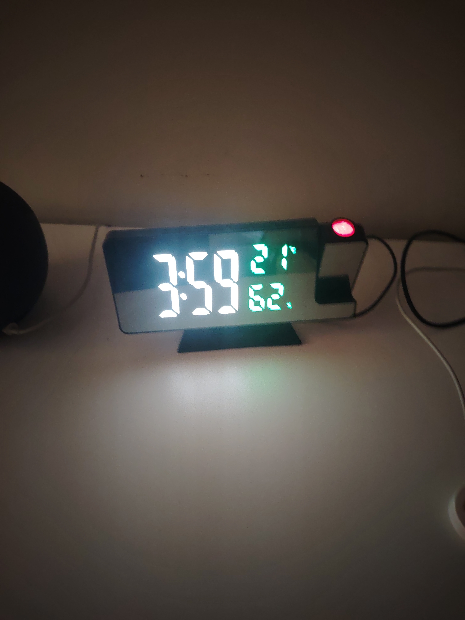 This image, taken in the city of Huntingdonshire, United Kingdom, depicts a digital alarm clock and associated electronic devices on a light-colored surface, likely a desk or bedside table. The central object is a modern digital alarm clock displaying the time "3:59" in large, bright white segmented digits. Above the right side of the time, smaller green segmented digits show "21°" (indicating temperature, likely Celsius), and below that, "62." (likely indicating humidity). The clock unit features a small, illuminated red circular component on its top right, which appears to be a projection lens. To the left of the clock, a dark, spherical electronic device, possibly a smart speaker, is partially visible. White and black cables are present on the surface to the right of the clock, with a small, round, light-colored object partially visible in the bottom right corner of the frame. The background consists of a plain, light brown or beige wall. The scene is illuminated by the clock's display and ambient light, suggesting an indoor, possibly nighttime or low-light, environment. No individuals are present in the image.