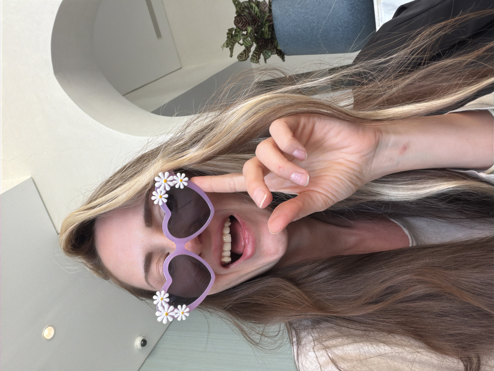 A vibrant, close-up portrait captures a young woman in an indoor setting, striking a playful pose with her head tilted, suggesting an upside-down or sideways perspective. She has long, flowing hair with shades of blonde and light brown, framing her face.
The woman is wearing distinctive purple, heart-shaped sunglasses adorned with small white daisy decorations on the top outer edges. Her mouth is wide open in what appears to be a cheerful laugh or enthusiastic smile, revealing her teeth and tongue. One hand is raised near her face, with fingers arranged in a peace or V-sign gesture, further emphasizing her playful mood. A faint mark or scar is visible on her left wrist in one of the close-up crops.
The background is bright and consists of light-colored walls or ceiling, indicating a well-lit interior, likely during the daytime. Visible elements in the background include a curved white architectural feature, a glimpse of a potted plant with dark green leaves in a blue container, and what appears to be a wall-mounted light or small camera. There is no visible text in the image. The overall impression conveyed is one of joy, casualness, and fun, captured in an indoor environment in Sharm El Sheikh, Egypt.