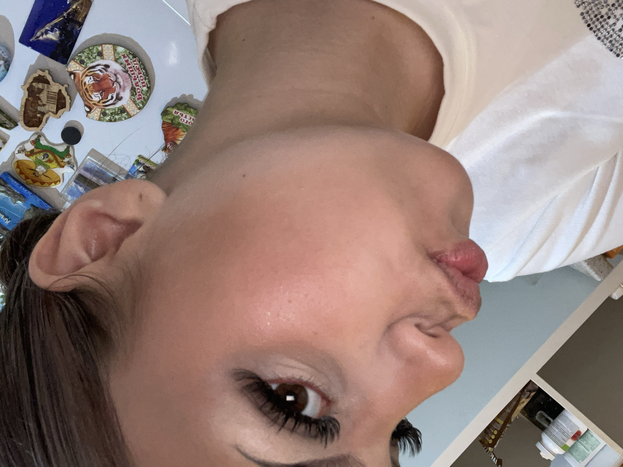 A close-up selfie captures a person, likely female, with dark hair, a light complexion, and brown eyes. Her face is prominent in the frame, tilted as if the photo was taken while lying down or at an angle. She is making a playful pouting or "kissing" face with her lips. Her eye features long, dark eyelashes, possibly false, and her skin appears clear. She is wearing a light-colored (possibly white or off-white) t-shirt.
The background reveals a surface adorned with several refrigerator magnets, suggesting an indoor setting like a kitchen or dining area. Notable magnets include a circular one with a tiger's head in the center, featuring Russian text that reads "БОЛЬШОЙ КИШМОН" (Bolshoy Kishmon). Other magnets visible are a blue rectangular one depicting an aircraft, a wooden magnet with a building design, and another irregularly shaped magnet with a cartoon-like crocodile. A partial Russian word "ОТО..." is visible on a green magnet. In the lower right corner, the edge of a shelf or cabinet is visible, containing bottles or containers. The lighting is soft and even, typical of an indoor environment. The overall mood conveyed by the subject's expression is playful or whimsical.