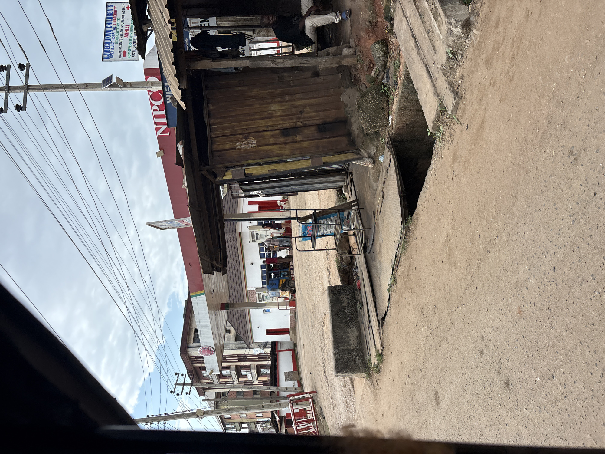 The image shows a street scene in Onitsha, Nigeria, focusing on a gas station and a small roadside stall. A prominent Nipco gas station with red and white branding dominates the background. In front of the gas station, a blue and yellow auto-rickshaw is parked, with several people standing nearby. To the right of the gas station, a rustic wooden stall with a corrugated metal roof is situated. Inside this stall, a man in a black shirt and light-colored pants is seated on a bench, looking towards the right. Various items are visible around the stall, including clothing hanging on a post and some shelves with items. The ground in the foreground is a dusty, unpaved road with a concrete structure and a ditch or open drain. In the far left of the image, a tall building with multiple floors and balconies is visible, suggesting an urban environment. The sky is overcast with scattered clouds.