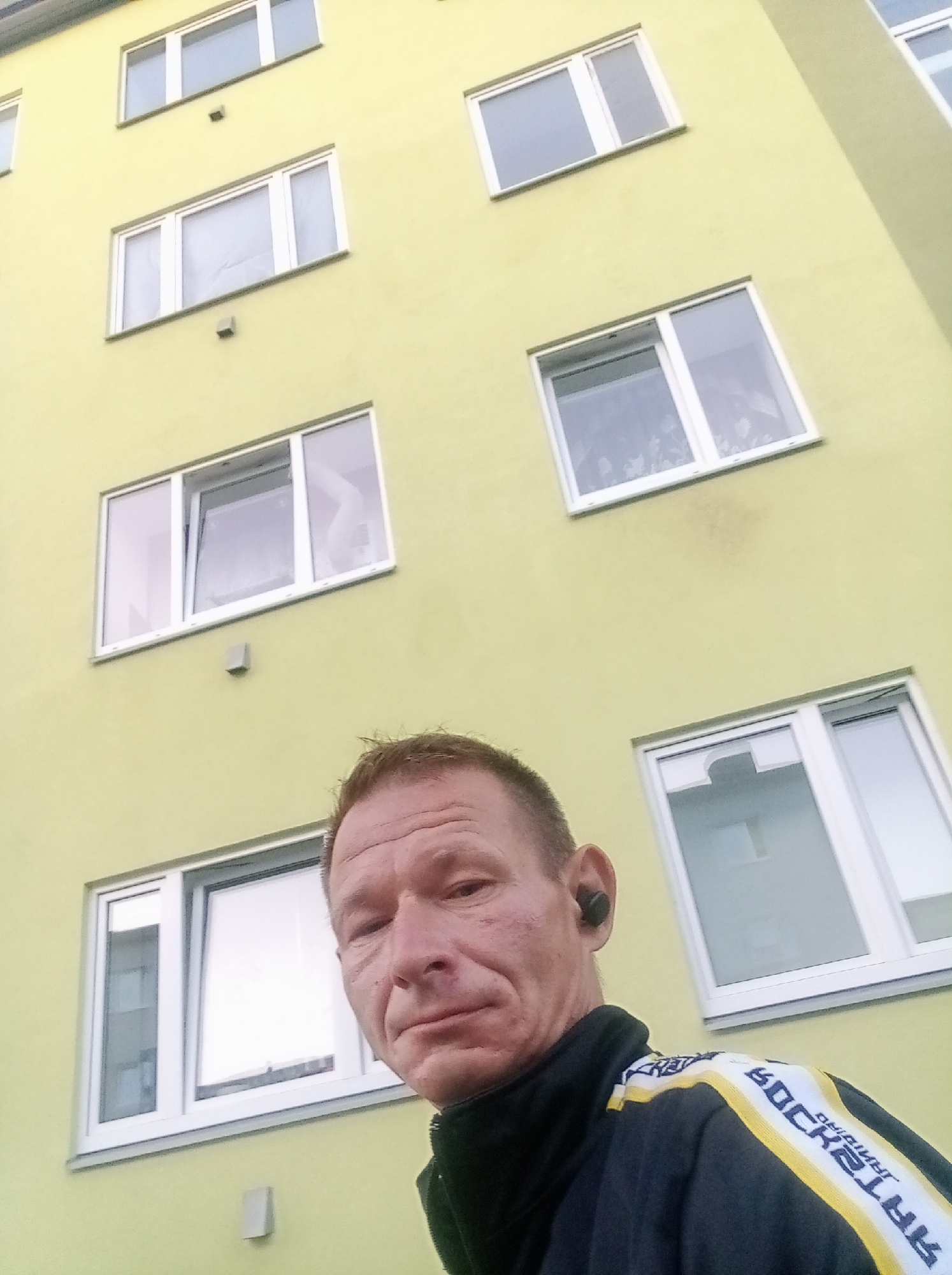 The image shows a man standing in front of a pale yellow building with multiple windows. The man is wearing a black jacket with white and yellow stripes on the sleeves and shoulder. He has dark hair and is wearing a black earbud in his right ear. The building appears to be a multi-story apartment building, with rows of evenly spaced windows. The windows are modern, with white frames and clear glass. Some of the windows have a slight reflection of the sky or surrounding environment. The building is located in the city of Tapa, Estonia. The perspective of the photo suggests the man is taking a selfie or is being photographed from a low angle. The overall environment appears to be an urban or suburban setting, given the architecture of the building.