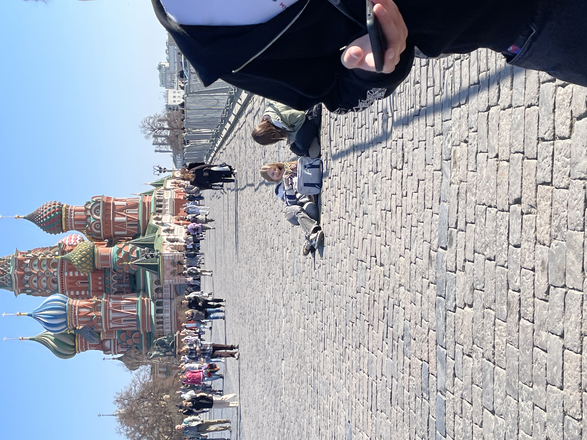 The image shows St. Basil's Cathedral in Moscow, Russia, viewed from Red Square. A large crowd of people is walking in front of the cathedral. Two young women are sitting on the cobblestone pavement in the foreground, possibly interacting. Part of another person holding a smartphone is visible at the top of the frame, potentially taking a photograph. The weather appears sunny and clear. Metal barriers are visible on the right side of the square.