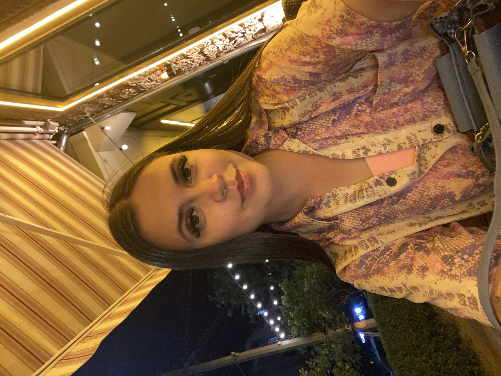 A young woman with long, dark hair is taking a selfie at night, sitting outdoors under a striped awning. She is wearing a patterned shirt with pink and yellow tones and a light pink top underneath. The background shows string lights and trees, suggesting an outdoor dining area or a street at night. There are illuminated architectural elements with golden trim and a large window reflecting the lights. The overall atmosphere is casual and illuminated by artificial light. The location is identified as the city of Derbent, Russia.