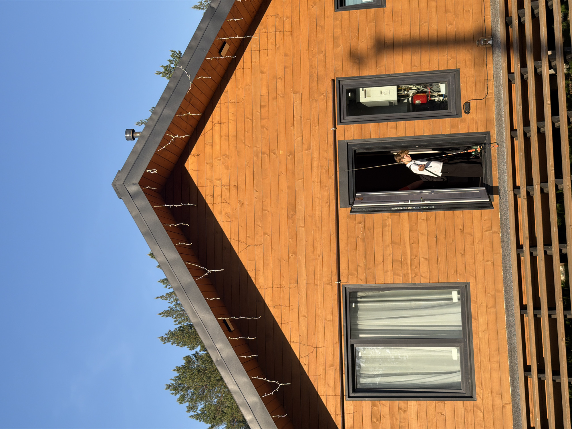 A medium shot of a brown wood-paneled home in Kamyennoye, Russia on a clear day with a man standing in an open window. The house has a dark gray roof angled downward to the left, decorated with sparse, small white lights. To the left of the house, trees with green foliage are visible against the clear blue sky.
The right side of the house features three vertically stacked rectangular windows with dark frames. The bottom window is covered by white curtains. The middle window is open, and a man is seen holding fishing rods. The top window shows a reflection of items inside the house, including what appears to be a white appliance with red and silver attachments. A narrow, dark slat structure runs vertically alongside the right edge of the house. The overall impression is of a quiet residential scene in daylight.