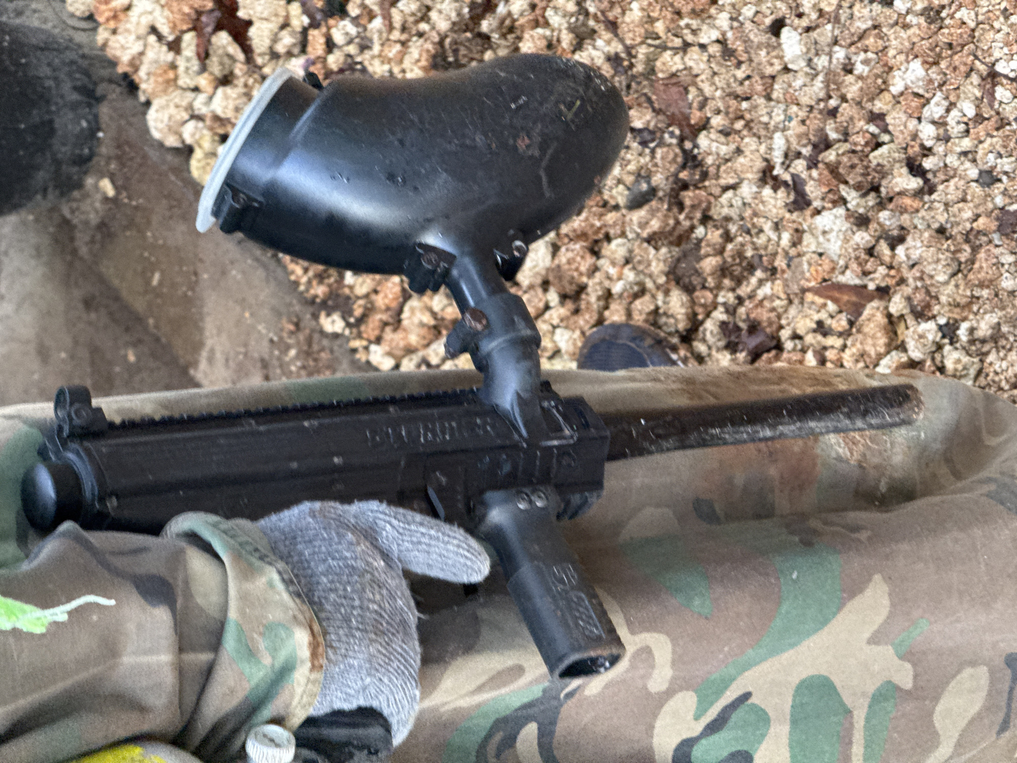 The image shows a black paintball marker resting on camouflage fabric. The marker has a large hopper attached to the top. A gloved hand is visible near the grip. The background includes small gravel or pebbles. The location is unspecified.
