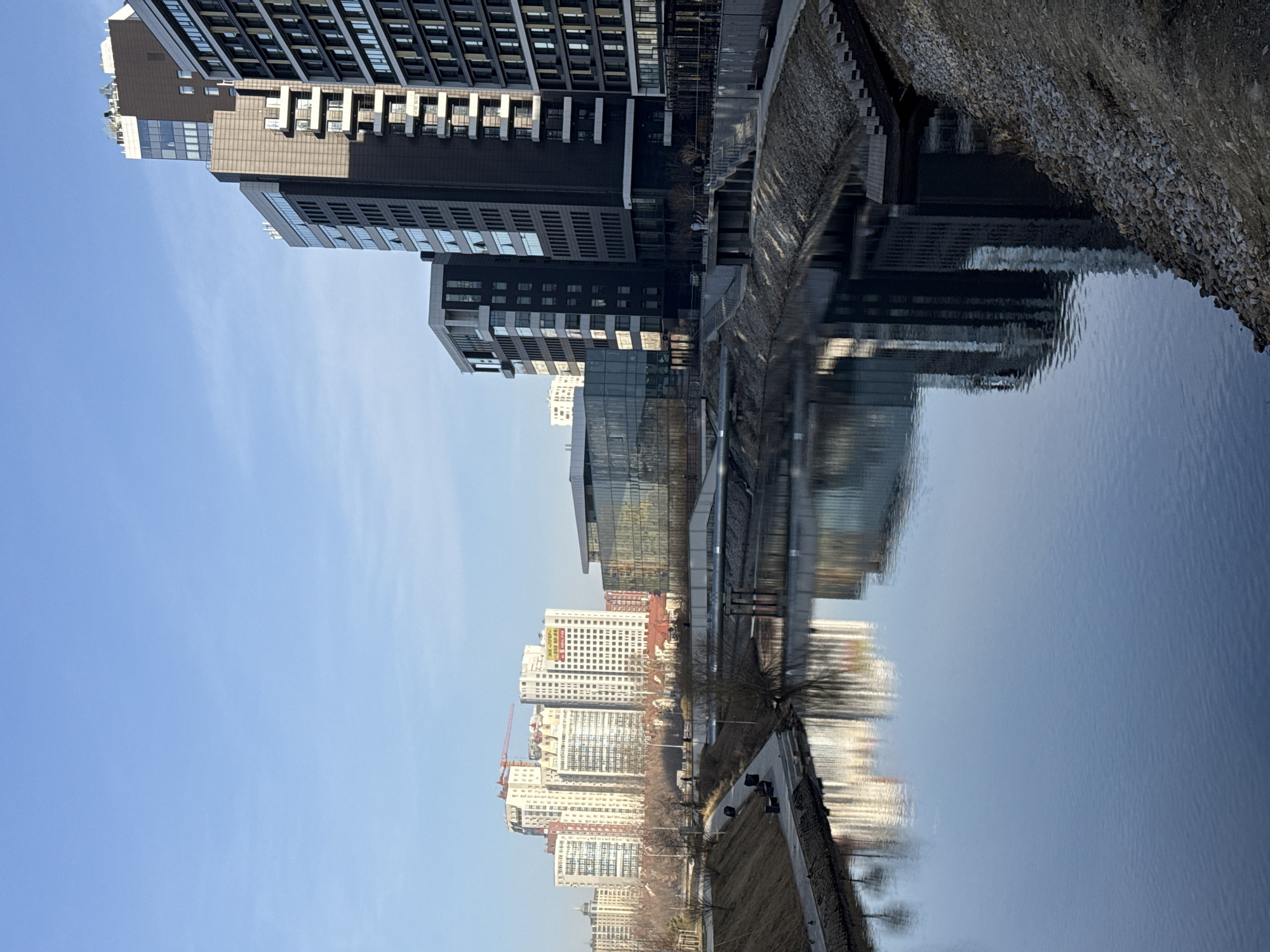 The image depicts a cityscape in Yekaterinburg, Russia, reflected in a body of water. Modern high-rise buildings with a mix of dark and light facades dominate the upper portion of the image, their reflection visible in the calm water. Further along the water's edge are lighter-colored residential buildings, also reflected in the water. The embankment is visible on both sides of the water, with pathways and vegetation. The sky is clear and blue, visible in the upper left portion of the image.