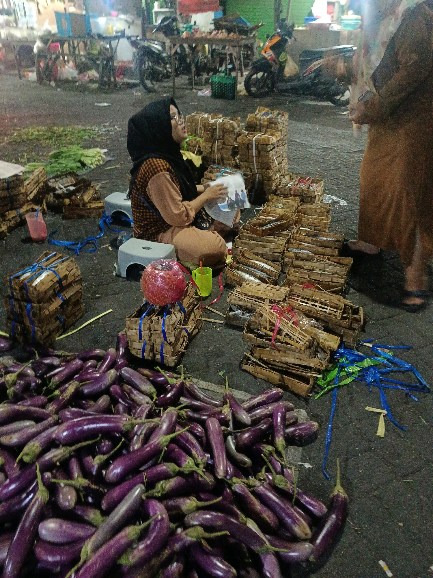A street market scene is depicted at night in Wonocolo, Indonesia. In the foreground, a large pile of fresh, purple eggplants lies on the ground. Behind the eggplants, a woman wearing a black hijab and glasses is seated on a small stool. She is looking at a booklet or magazine. To her right are several stacks of small, woven baskets filled with what appear to be fish. The baskets are tied with blue and red ribbons. Further in the background, two motorcycles are parked, and market stalls with goods are visible. Another person, partially visible on the right side of the frame, is standing and wearing an orange dress. The ground is dark asphalt, and there are a few pieces of plastic sheeting and other debris scattered around. The overall lighting suggests artificial illumination from the market stalls.
