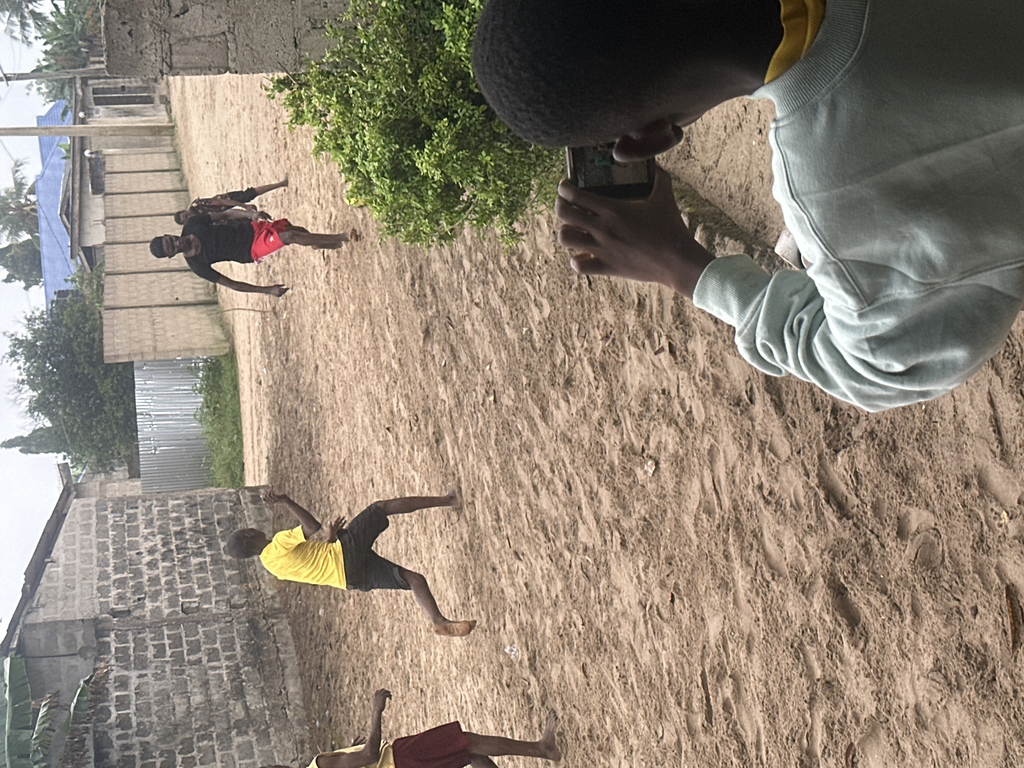 In Badagry, Nigeria, an outdoor scene unfolds with a group of young boys playing soccer in a sandy, unpaved area. The environment is characterized by dirt paths, some sparse vegetation, and various types of building structures, including cinderblock walls and corrugated metal fences. The sky appears overcast, suggesting potentially cloudy weather.
In the foreground, a young boy, viewed from the back, is holding a smartphone in landscape orientation, actively recording the events. He is wearing a light blue sweatshirt over a yellow t-shirt.
Further into the scene, three other boys are engaged in the soccer activity. One boy, wearing a bright yellow t-shirt and dark shorts, is mid-run, his body angled as if kicking or about to kick a ball. Another boy, in a black t-shirt and red shorts, is also running, with one leg extended, suggesting he is actively participating in the game. Behind him, a third boy, wearing a patterned short-sleeved shirt and dark shorts, is also running. The sandy ground is marked with footprints, evidence of their movement. The overall atmosphere suggests a casual, spontaneous game among children.