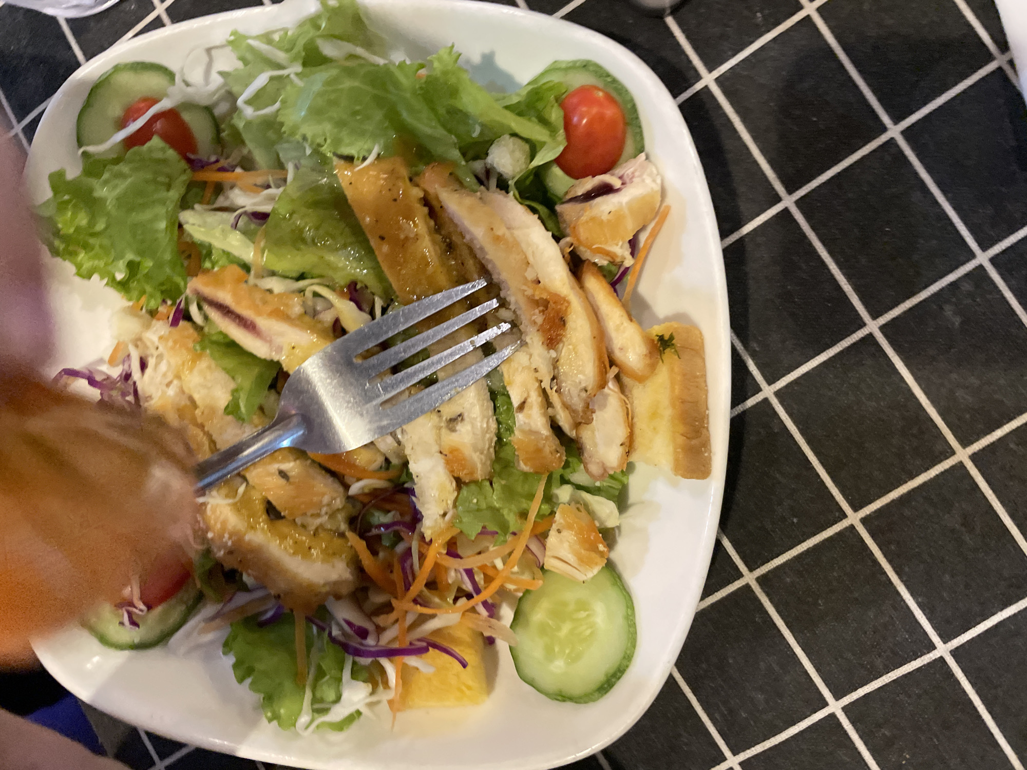 A slightly high-angle, full-shot depicts a white, square plate with rounded corners, positioned on a black-and-white checkered tablecloth. The plate contains a salad with mixed greens, sliced cucumbers, cherry tomatoes, shredded carrots, purple cabbage, pieces of orange, sliced grilled chicken, and croutons. A silver fork is visible, piercing a slice of chicken. A blurred portion of an arm and part of a shirt are visible at the lower left of the image. The setting suggests an indoor dining environment. The location, Bắc Nha Trang, Vietnam, is provided, though not visually verifiable.