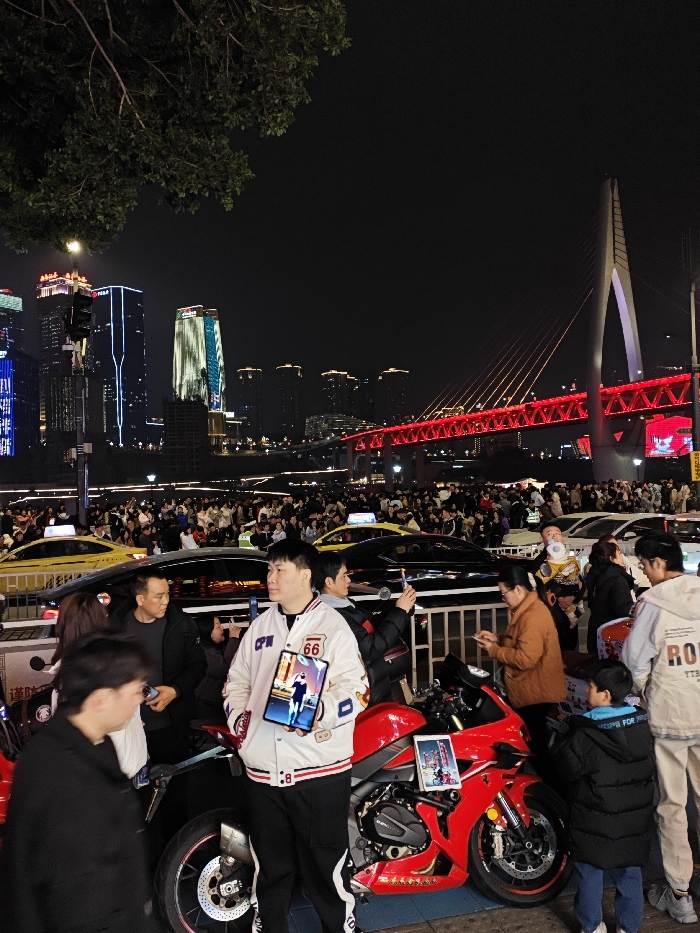 This image depicts a crowded nighttime urban scene within the Yuzhong District of Chongqing, China. In the foreground, a young East Asian male, approximately 20-30 years old, stands centrally. He wears a white varsity-style jacket featuring "CPW" and "66" patches, black athletic pants with white side stripes, and dark shoes. He holds a tablet in his left hand, displaying an image of a stylized figure. Immediately to his right, a red sport motorcycle is parked on the pavement; a second tablet, also displaying an image, is mounted on its fuel tank.
To the left of the primary subject, another individual is partially visible. Behind him, a man in a dark jacket is visible. To the right of the motorcycle, an East Asian woman in a brown jacket is looking downwards, and further right, a man in a light hoodie with "ROUTE" text stands near a child in a black puffer jacket. A dense crowd of pedestrians fills the middle ground, extending across what appears to be a street or plaza. Multiple individuals in the crowd are holding smartphones, some appearing to be recording or photographing the scene. Yellow taxis and dark-colored sedans are interspersed among the crowd, suggesting slow-moving traffic or parked vehicles. One person in the mid-ground holds a baby equipped with a distinctive white head support.
The background showcases the illuminated city skyline of Yuzhong District, featuring several tall buildings with distinctive light patterns. Dominating the right side of the background is the Dongshuimen Bridge, its main truss structure glowing bright red, while its prominent cable-stayed pylon is illuminated in white. The dark foliage of a tree is visible in the upper left corner, and a traffic light pole stands on the left. The scene is characterized by extensive artificial lighting from buildings, streetlights, and vehicle lights, indicating a bustling night atmosphere. Metal barriers separate the immediate foreground from the main pedestrian and vehicle areas.