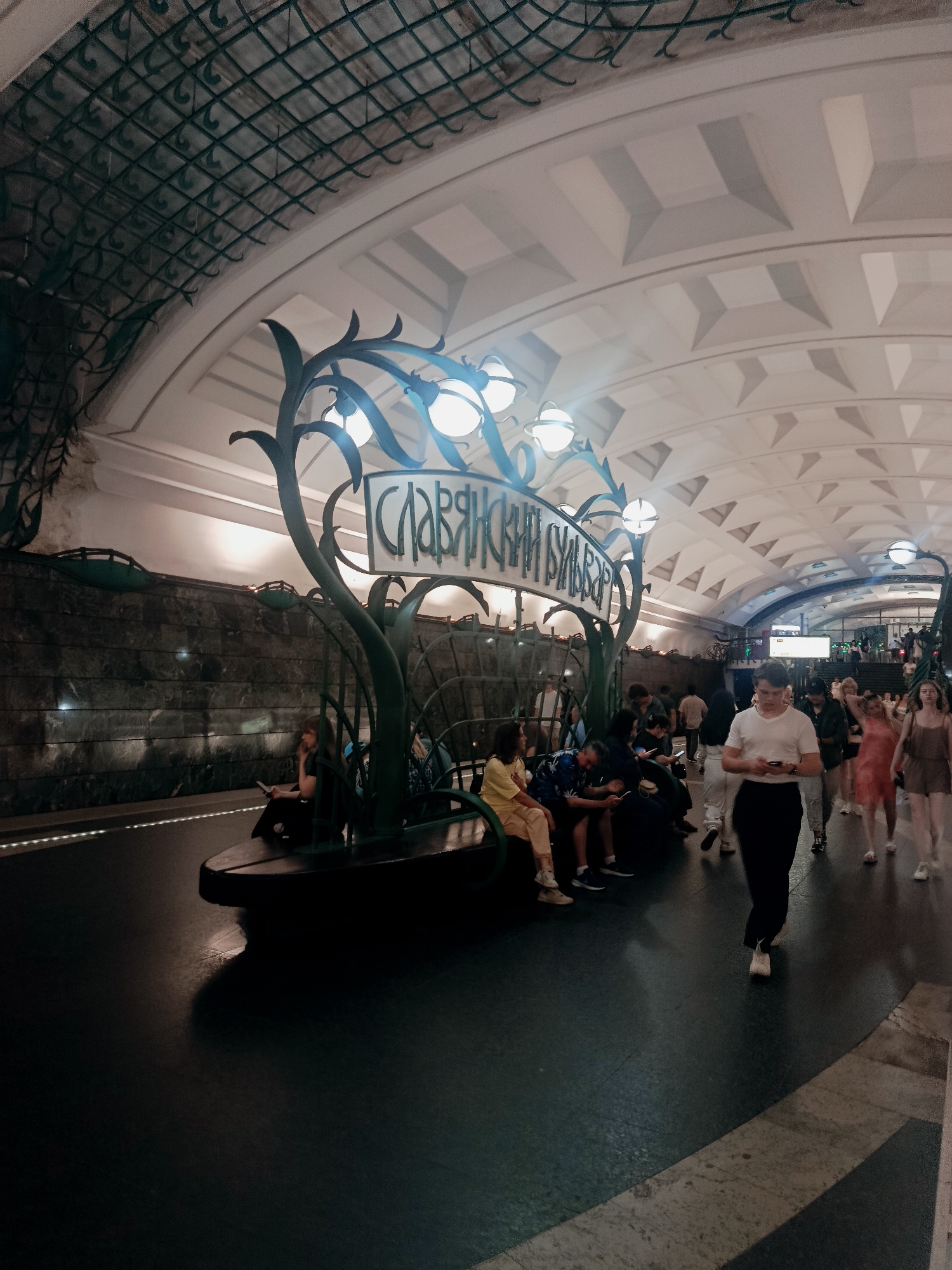 The image captures a busy scene inside the Slavic Boulevard metro station in Moscow, Russia. The station features an arched ceiling with a distinctive geometric pattern and a decorative metal lattice overhead. In the foreground, a stylized metal structure resembling a plant, with multiple lights, is a prominent feature. A bench is integrated into this structure, and several people are seated on it, some looking at their phones. A group of people are walking along the platform, which is dark and reflective. The station walls are made of dark, textured stone. The overall impression is that of a well-lit, architecturally interesting, and active public transport hub.