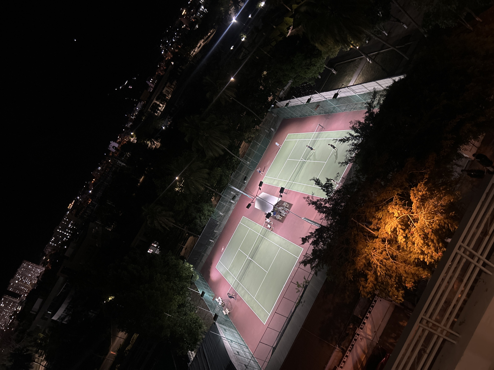 The image shows two illuminated tennis courts at night in Bac Nha Trang Ward, Vietnam. The courts are green with white lines, surrounded by a reddish-brown border. There are several people on the courts, appearing to be playing tennis. A net divides each court in half. In the background, a row of city buildings with lights is visible. Trees surround the tennis courts. A brightly lit tree occupies the lower right corner of the image, obscuring some of the view. A white metal railing is also visible in the lower right corner. A light pole illuminates the court in the center. There are chairs near the sidelines of the courts.