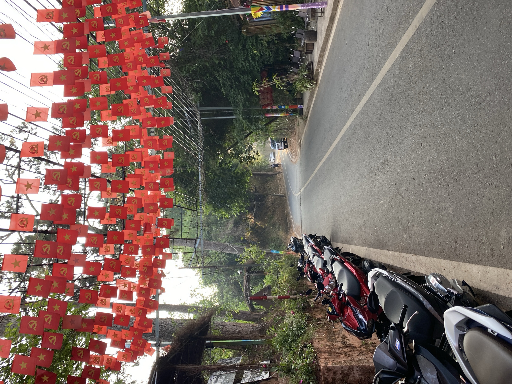 The image shows a street scene in Da Lat, Vietnam. Red flags, each bearing a gold star and hammer and sickle emblem, are suspended above the road. Motorbikes are parked along the side of the road. The road itself is asphalt, with a white dividing line. Vegetation is visible on either side of the road. A white vehicle is faintly visible in the distance.